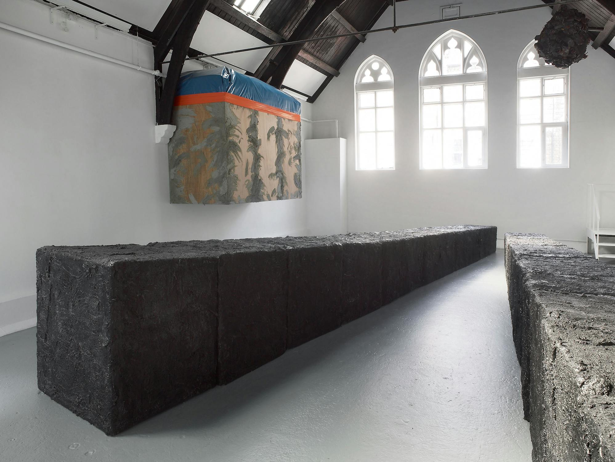 A modern gallery with high, dark wooden beams and white walls displays a large, rectangular, dark textured sculpture or installation. The structure forms a winding path or barrier, contrasting starkly with the clean, minimal space.