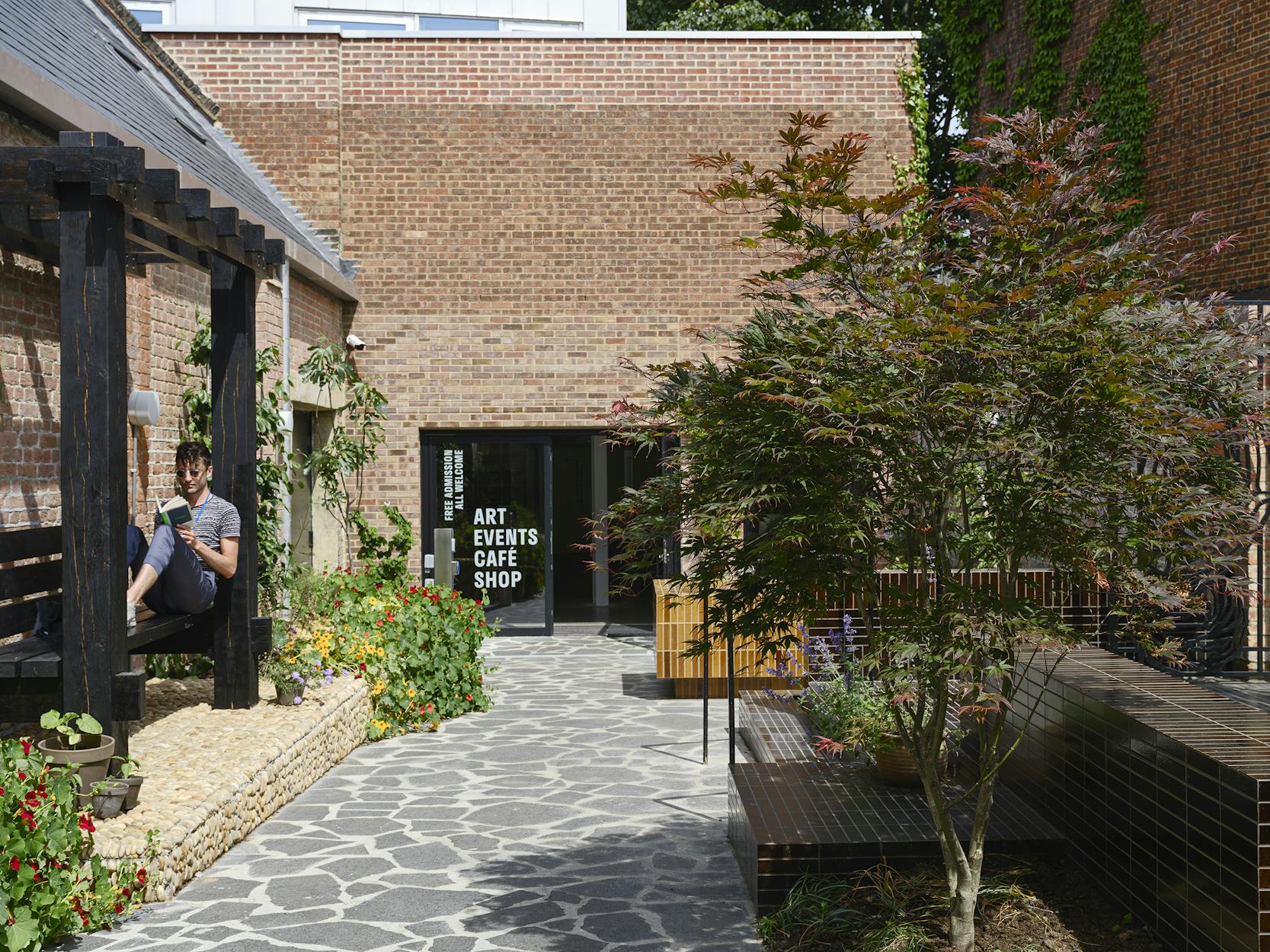 A courtyard with stone paving, plants and benches. A person sits on a pergola reading. In the background, a brick building entrance displays signs for art, events, a café and a shop.