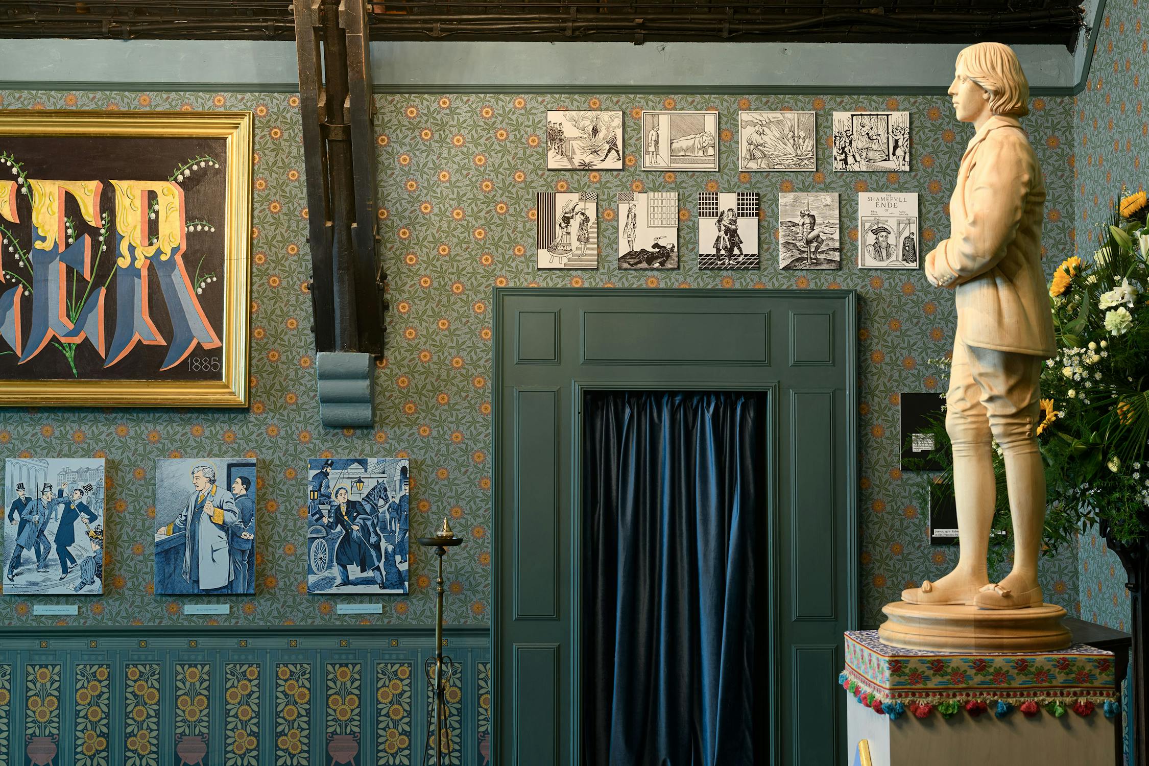 A room with ornate floral wallpaper features a white statue of a man in period clothing, blue curtains, comic-style artwork on the walls, and a colourful framed painting. Flowers in a vase are placed behind the statue on the right side.