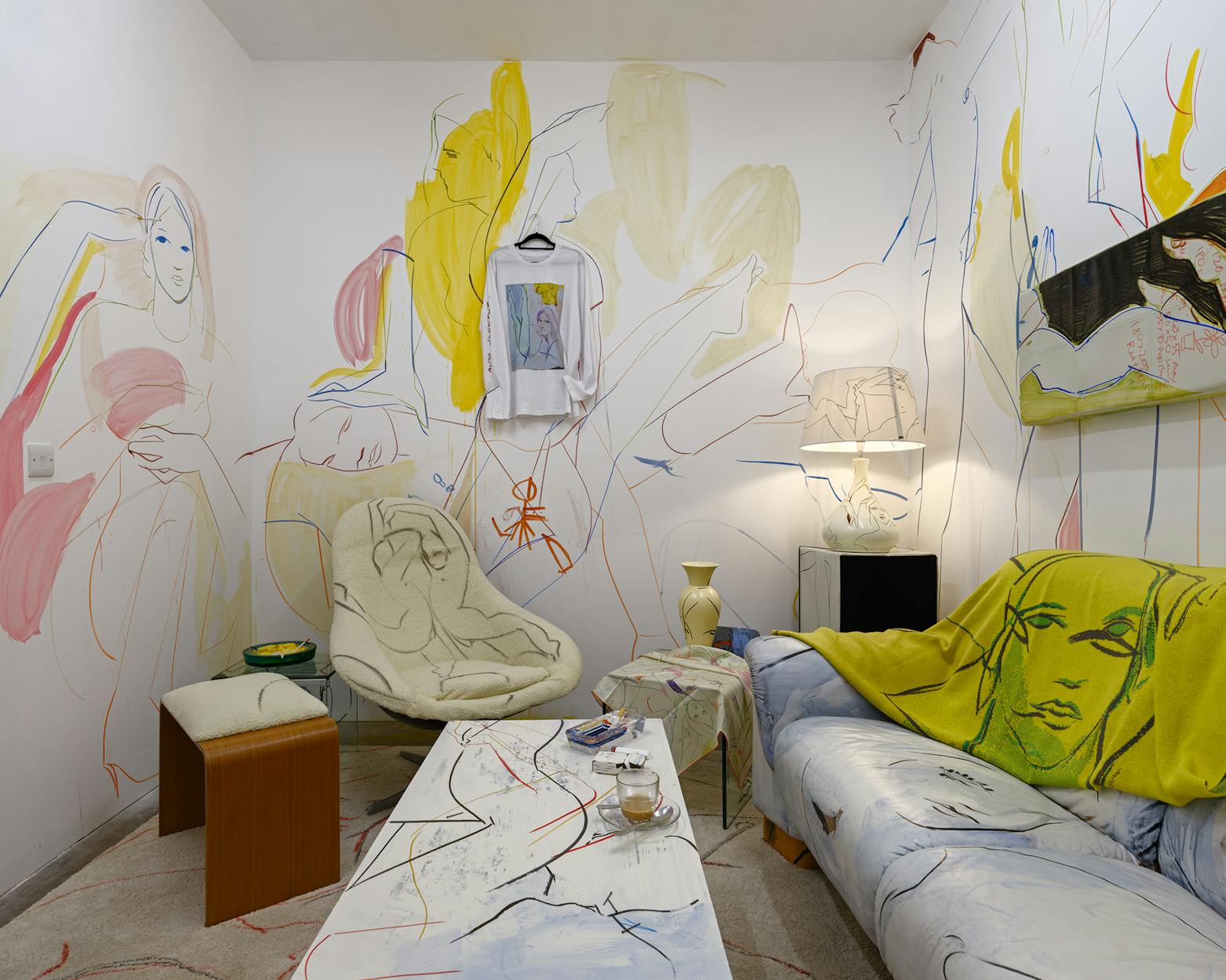 A living room installation with a modern, abstract line and colour mural covering the walls and furniture. The room has a plush chair, a sofa, a coffee table, and unique decor items, creating an artistic, creative atmosphere.