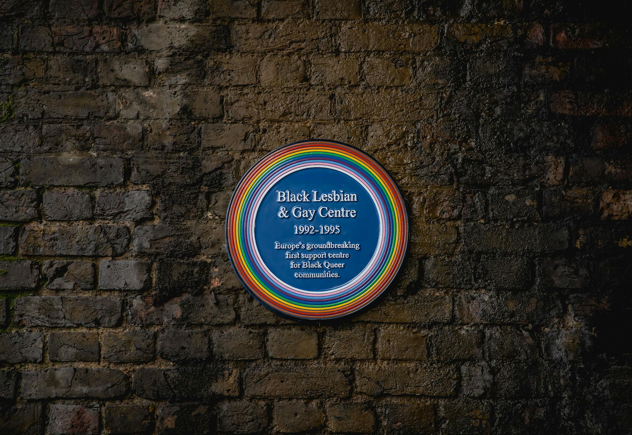 A circular wall plaque with a rainbow trim mounted on a brick wall.