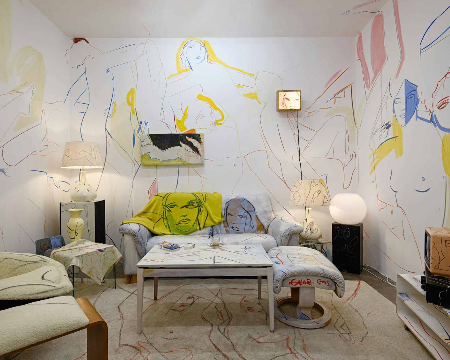 A living room installation by France-Lise McGurn with an abstract line mural of reclining figures covering the walls, furniture, and décor. The space features a couch, table, lamps, and colourful throws, all decorated with similar artistic designs.