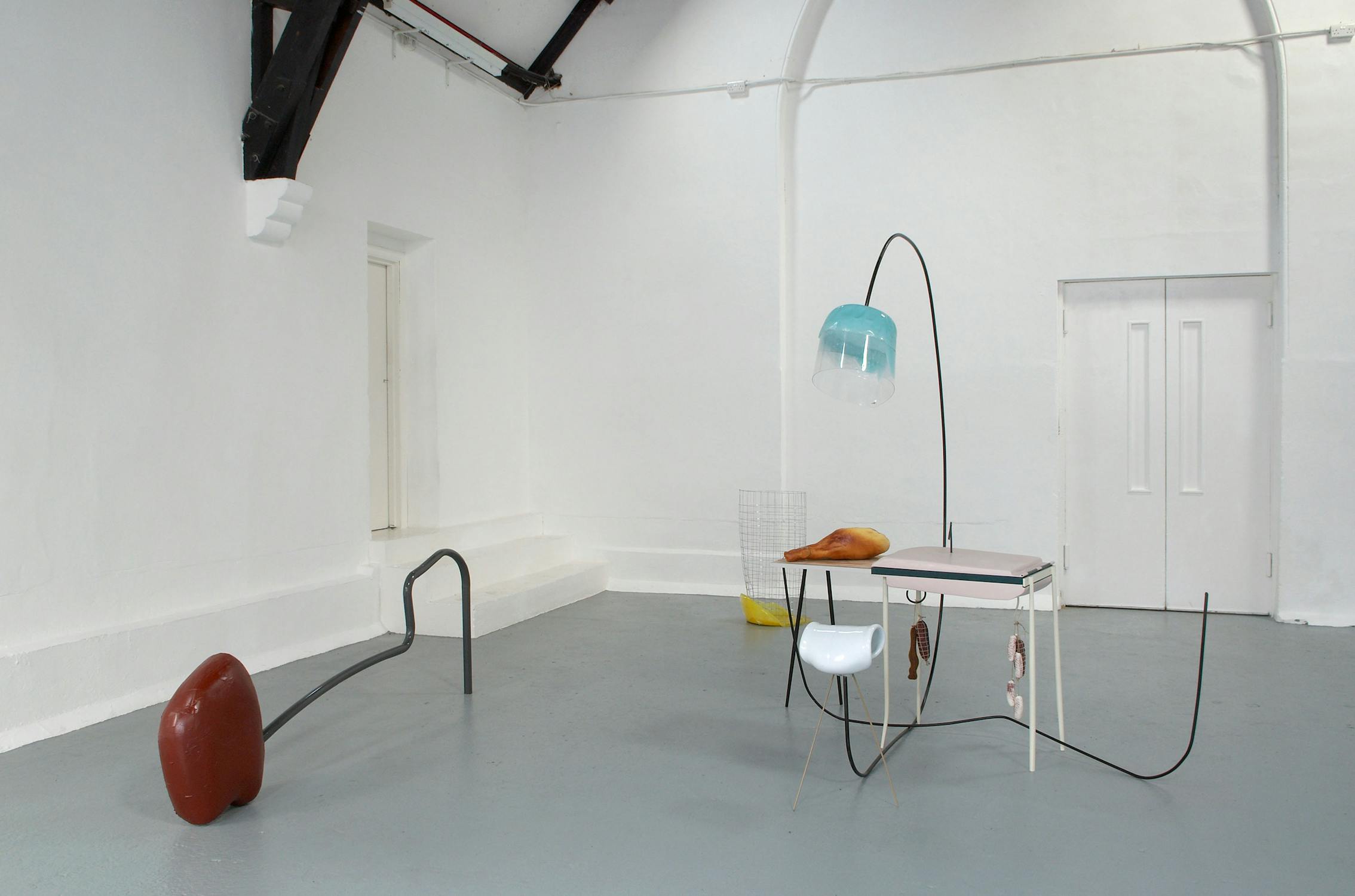 A minimalist art installation in a gallery with abstract sculptures. Objects include a red half-sphere, a curved black bar, a two-tiered table with a blue lamp, and a wire bin-like structure with a yellow substance at the bottom. The room is painted white and grey.