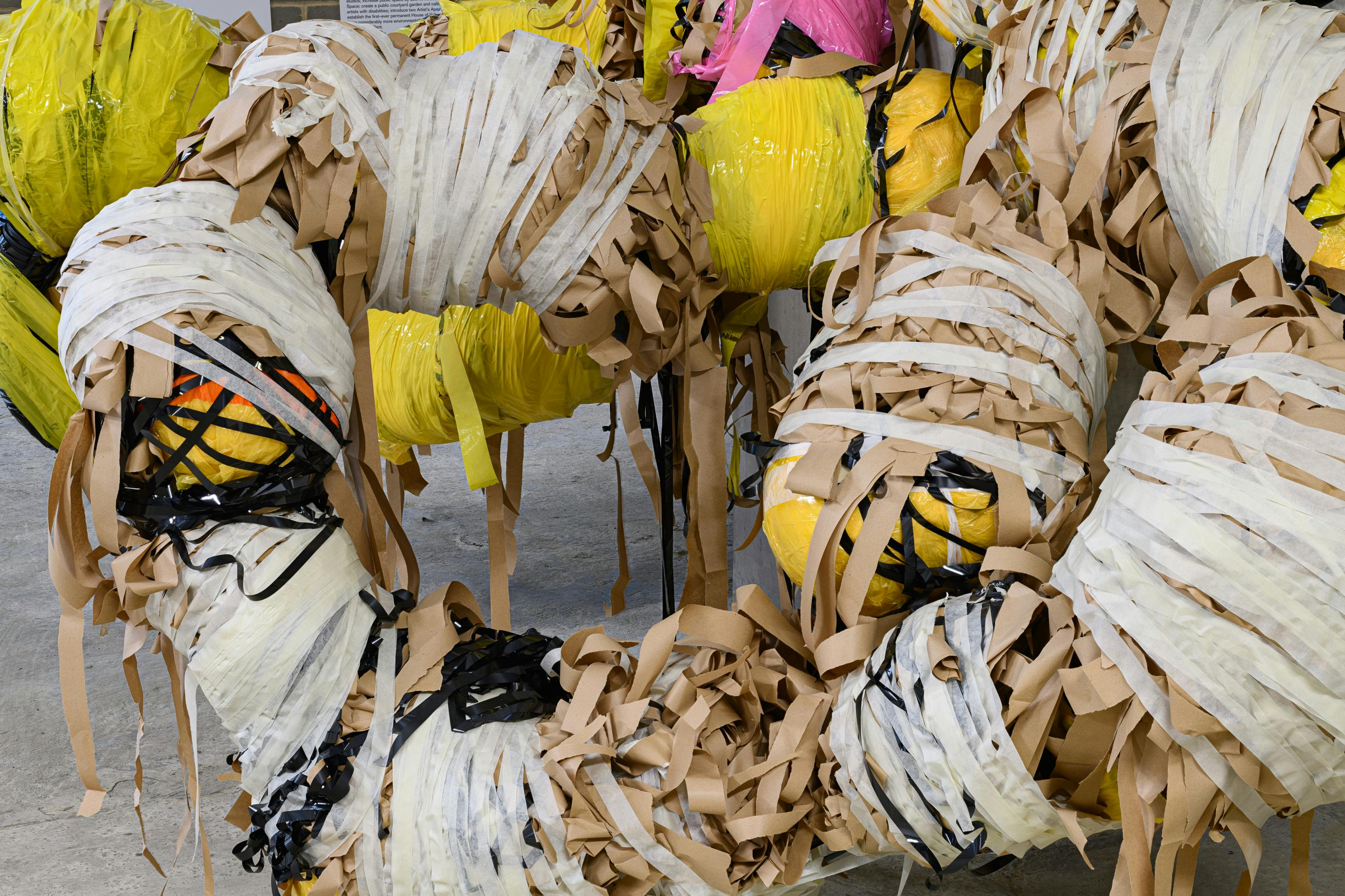 Bundles of yellow and pink objects tightly wrapped with layers of brown, white, and black tape or strips, creating a textured and chaotic appearance against a plain grey floor background.