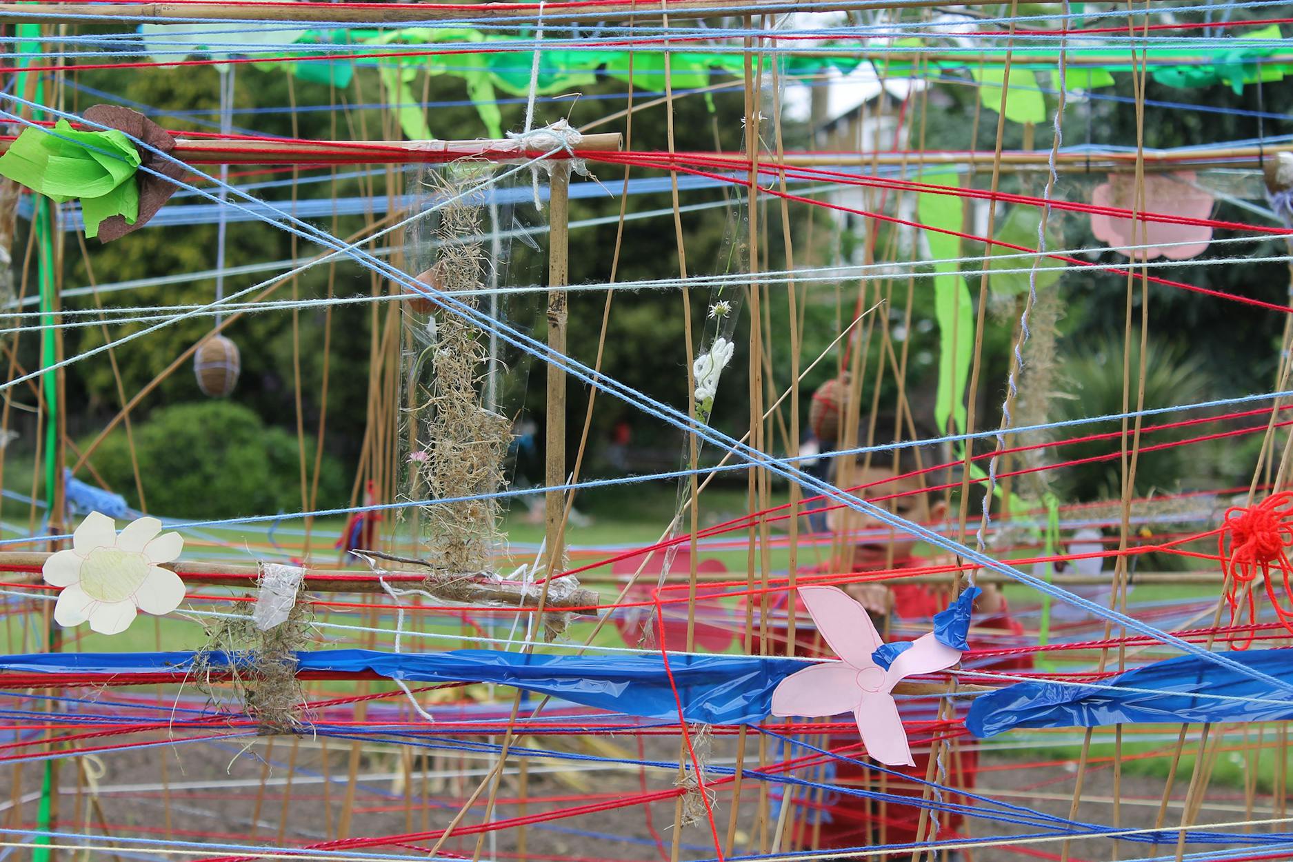 Close-up of an abstract outdoor art installation with colorful strings, fabric, and paper flowers interwoven. The setup features red, blue, and yellow strands with natural materials, set against a green park background.