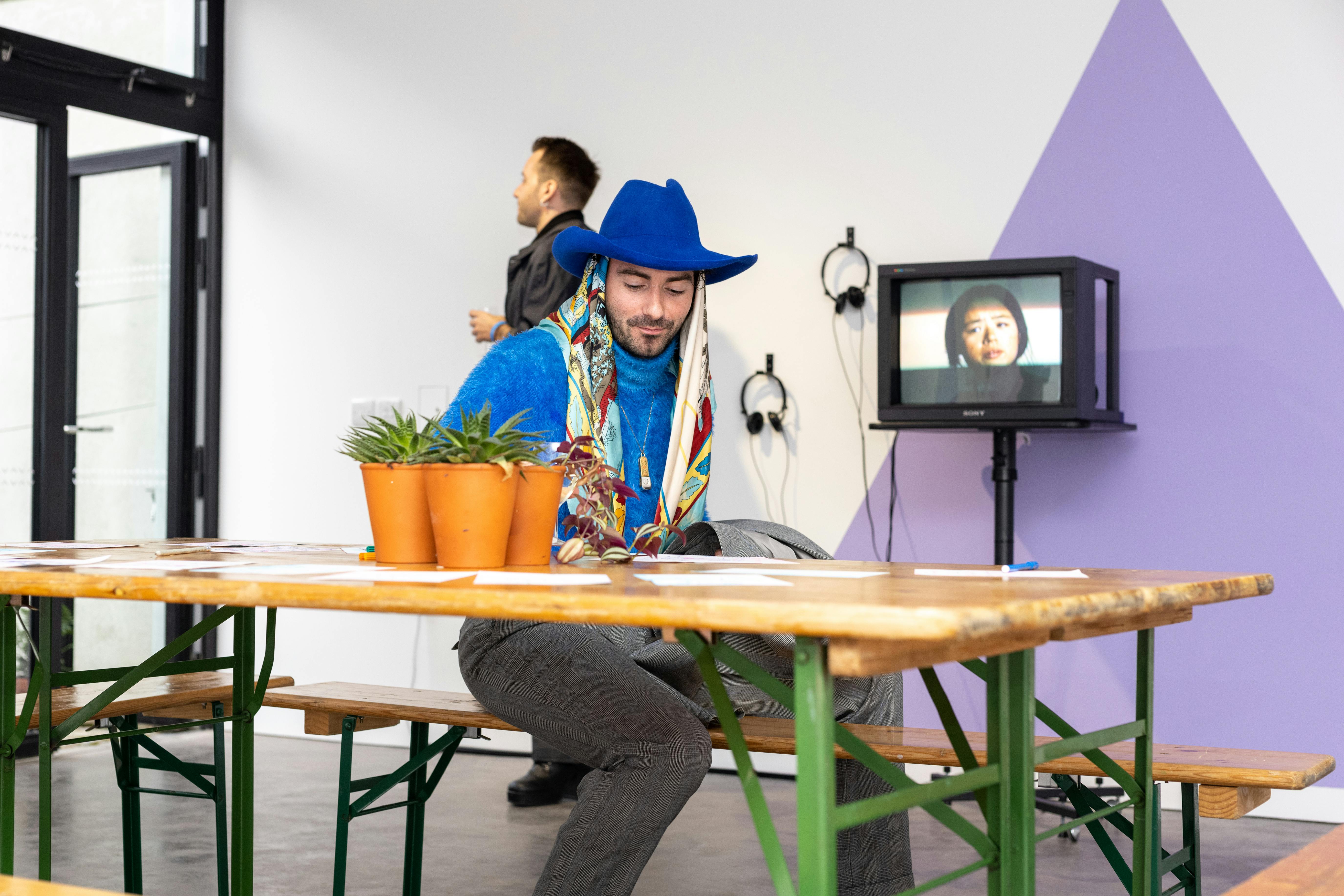 A photograph of a man sitting at a folding table. He wears a blue hate and jumper with a colourful silk scarf. In the background there is a TV on a wheeled stand, and a pair of headphones hung on the wall.