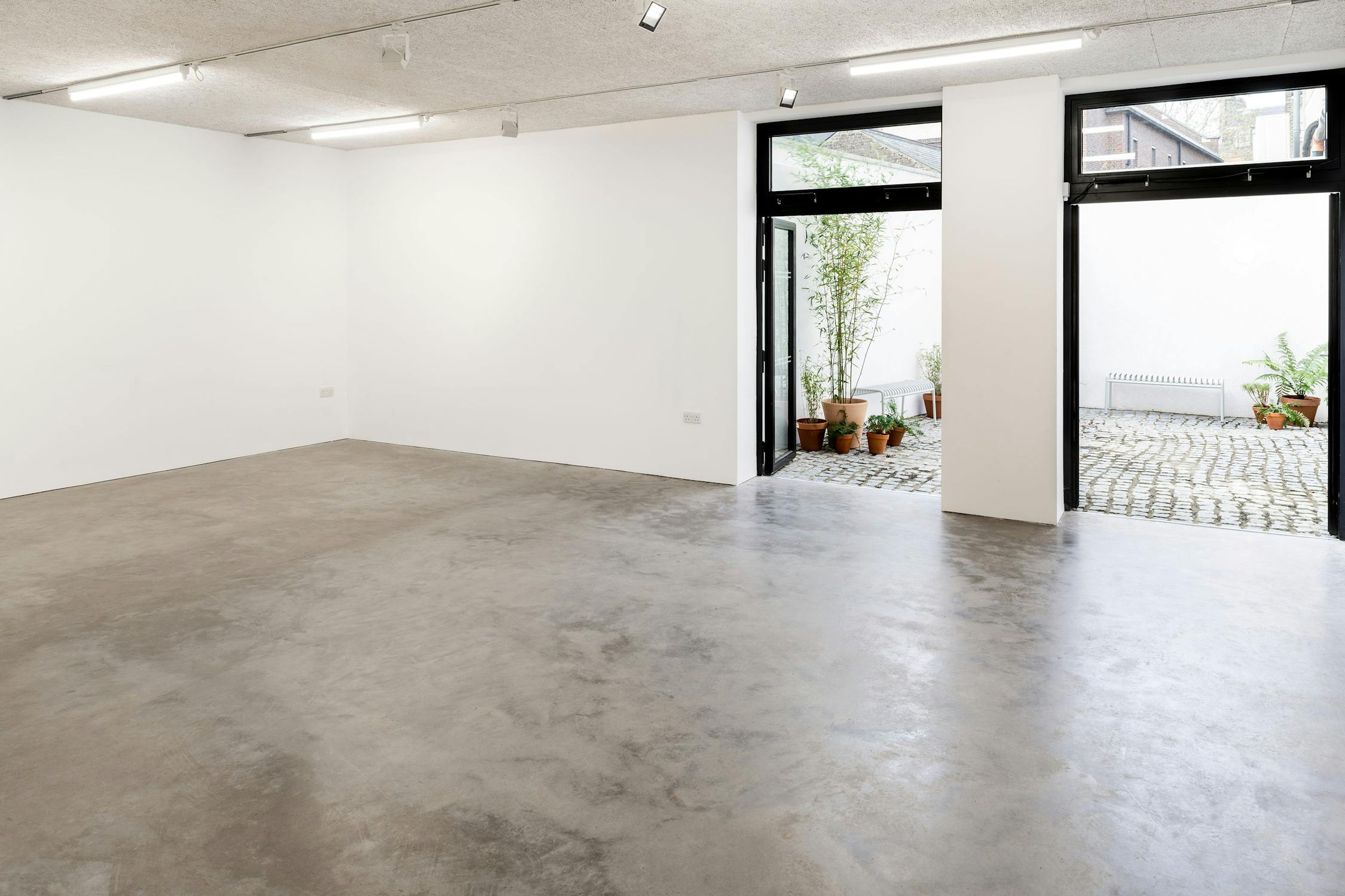 a large empty room with white walls, and a polished concrete floor. Two large doors open onto a paved courtyard