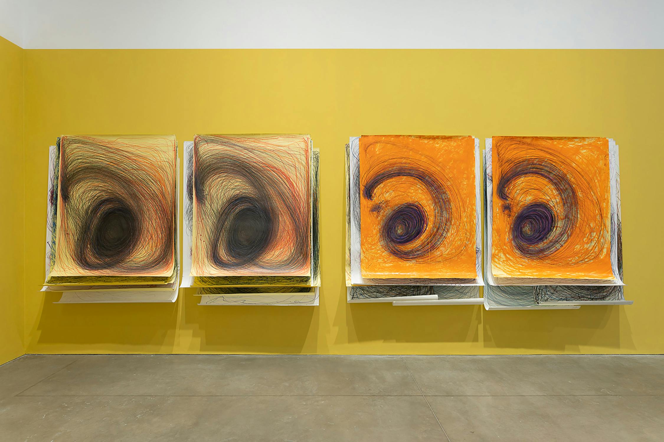 Four abstract artworks displayed on yellow walls, each featuring large swirling patterns in dark and orange hues. They are mounted individually, with light illuminating their textured surfaces.