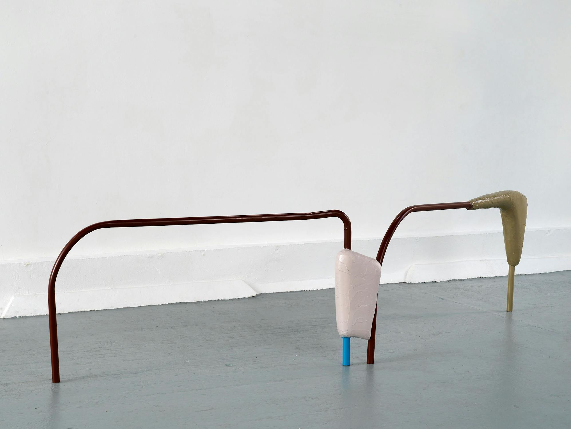 A minimalist art installation with two brown metal rods extending from the floor, each topped with smooth, abstract forms—one beige and one light grey. The setup stands against a white wall on a light grey floor.