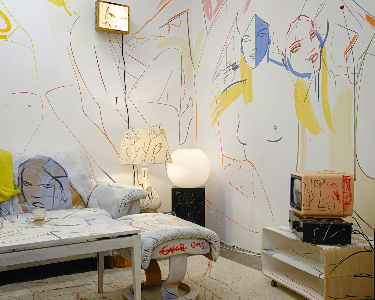 Art-filled living room with abstract line drawings of faces and bodies on the walls, furniture, and decor. A white chair, table with a glass, unique lamps, and a TV are visible in the France-Lise Mcgurn-designed space.