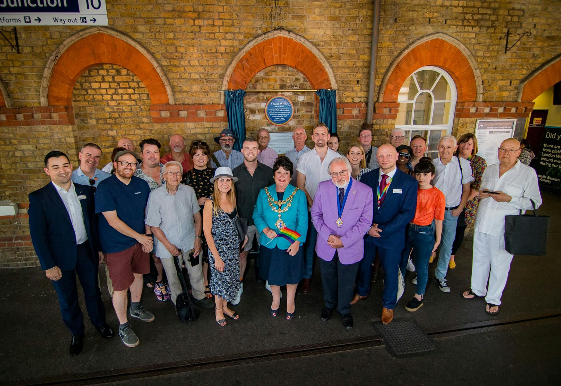 A group of around 25 people, some in formal attire, pose together inside a train station near a brick wall with arched windows and a blue plaque with rainbow edge honouring Oscar Wilde. Signs for platforms and exits are visible above them.