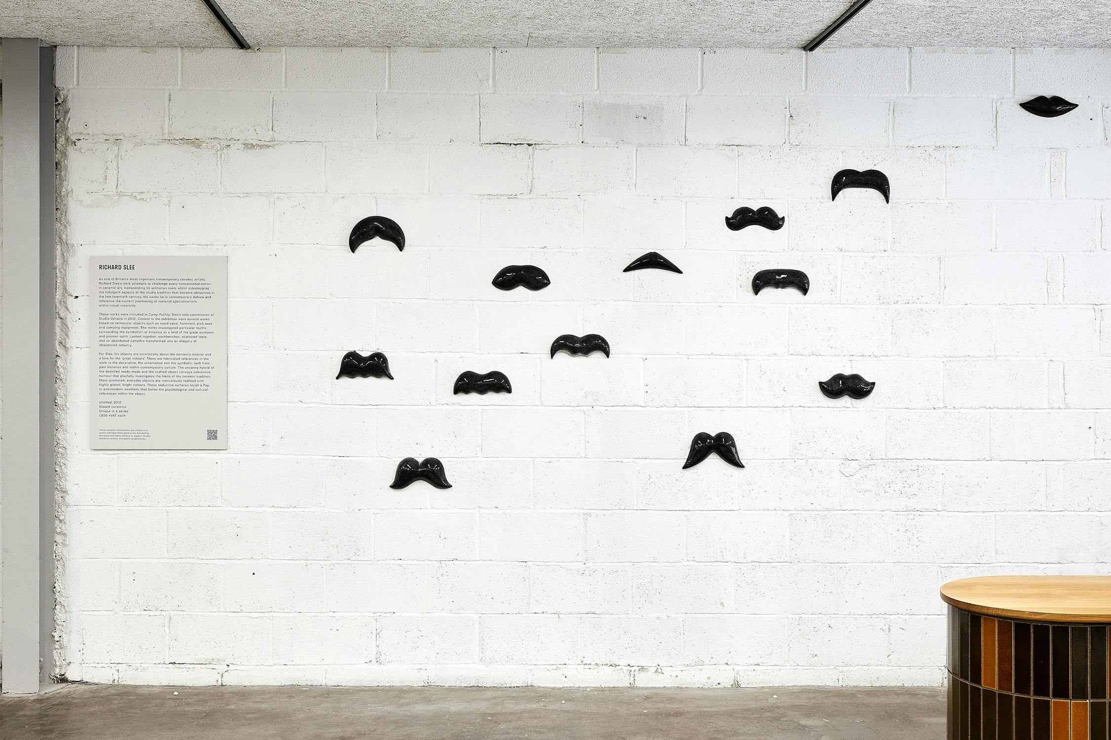 a photograph of an installation of sculptures that resemble moustaches. They are glazed in grey and are arranged on a wall. In the foreground there is a round table with tiled base.
