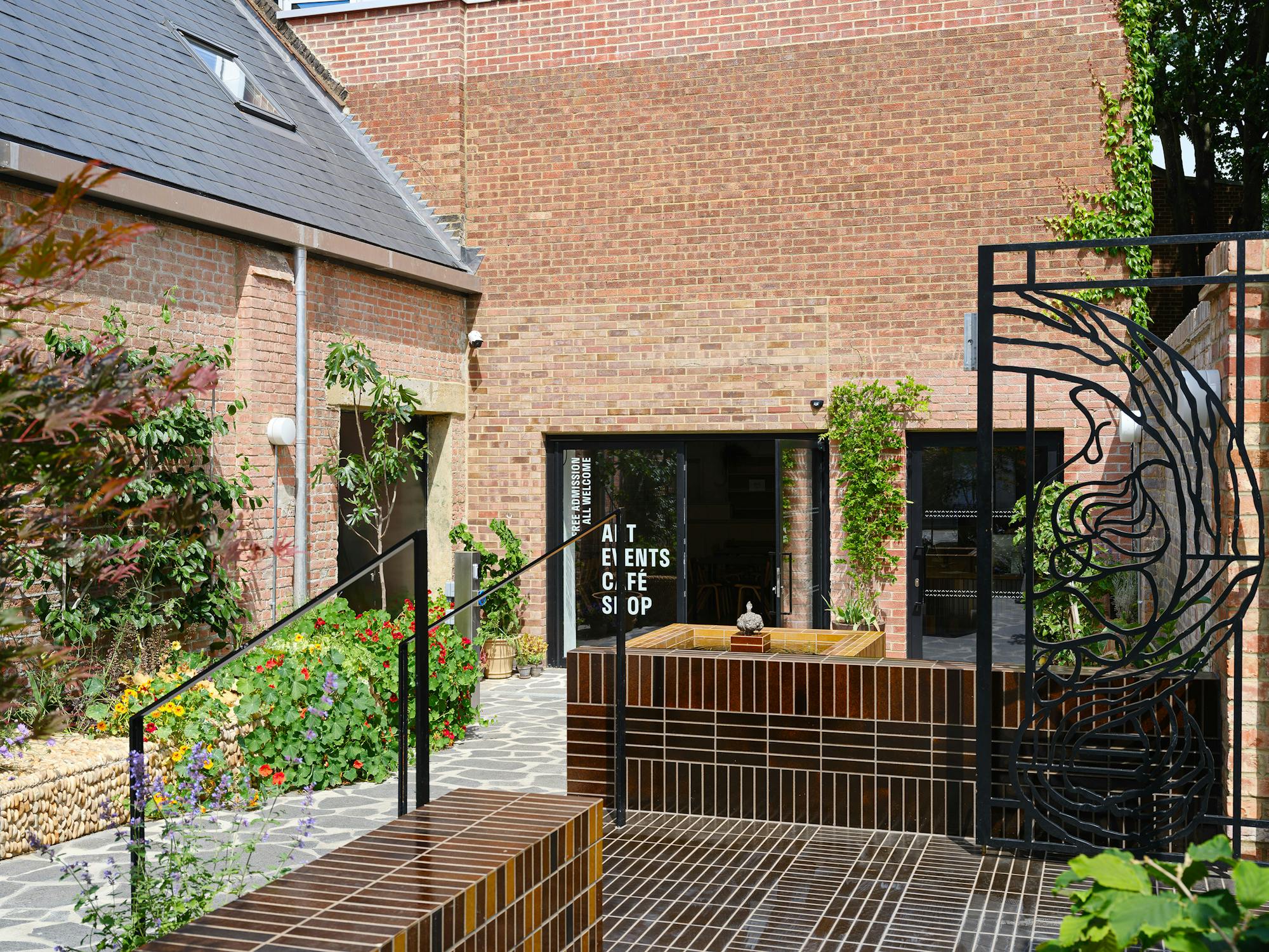 A brick building with a black modern gate featuring artistic designs. The entrance is adorned with green plants and colourful flowers. The courtyard includes brown tiled seating areas and a stone pathway leading to the door.