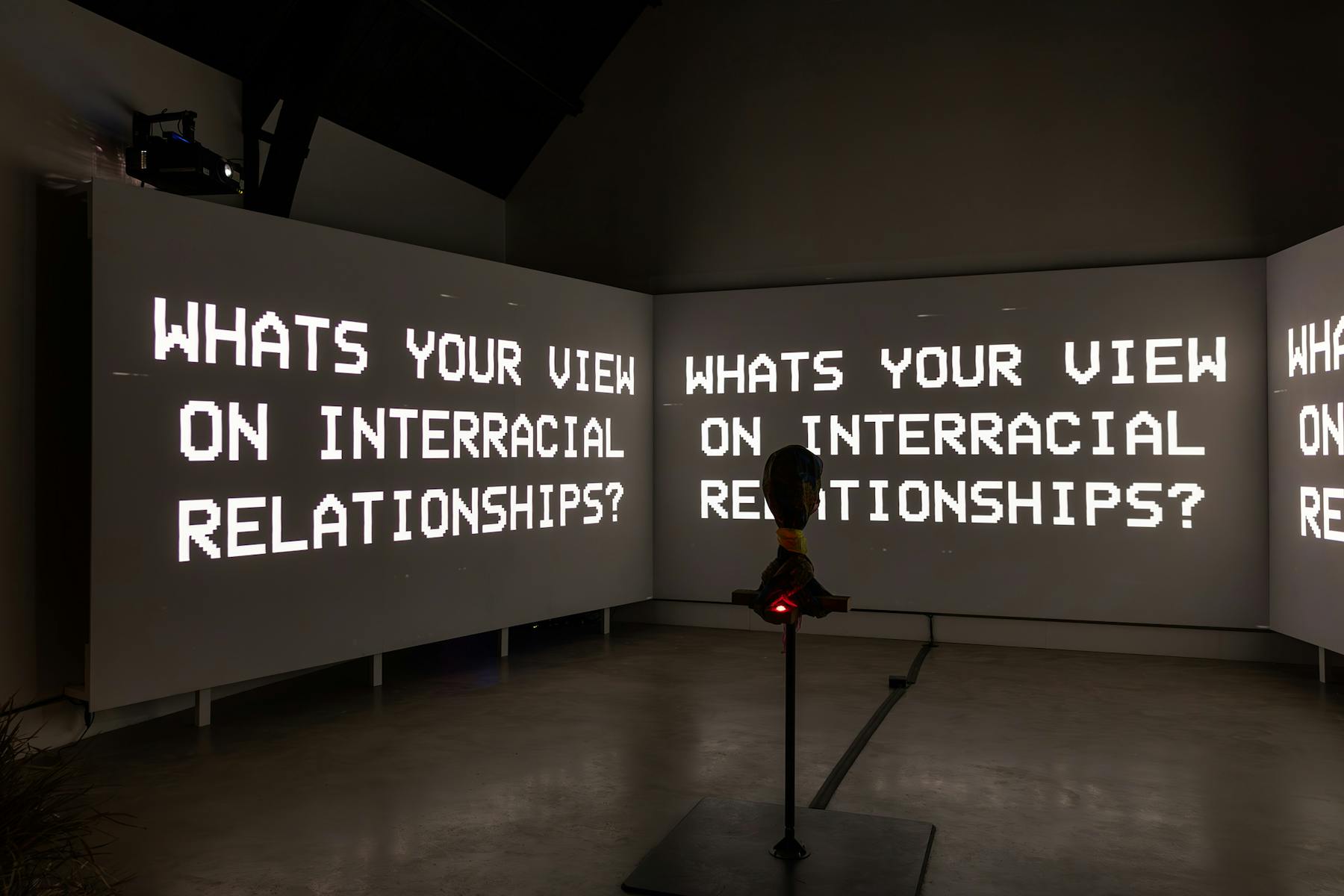 a photograph of three screens that are projected with black and white text. There is text that reads 'WHATS YOUR VIEW ON INTERRACIAL RELATIONSHIPS'. In the foreground there is a hooded object on a stand. 