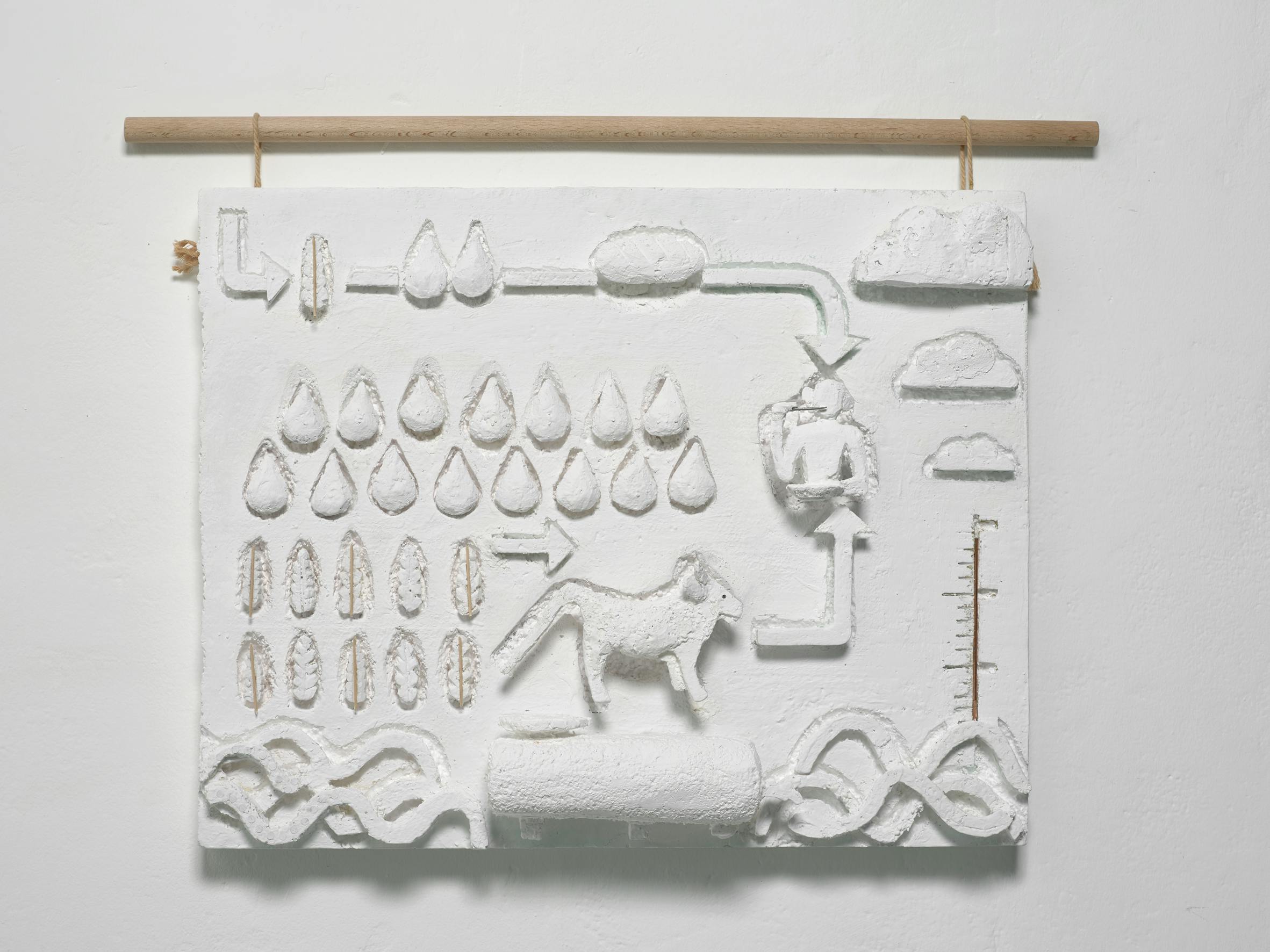 A white textured relief artwork hangs on a wall from a wooden rod. It features abstract shapes: a central animal (possibly a fox or wolf), water droplets, arrows, pipes, clouds, and waves, suggesting a scientific or ecological diagramme.