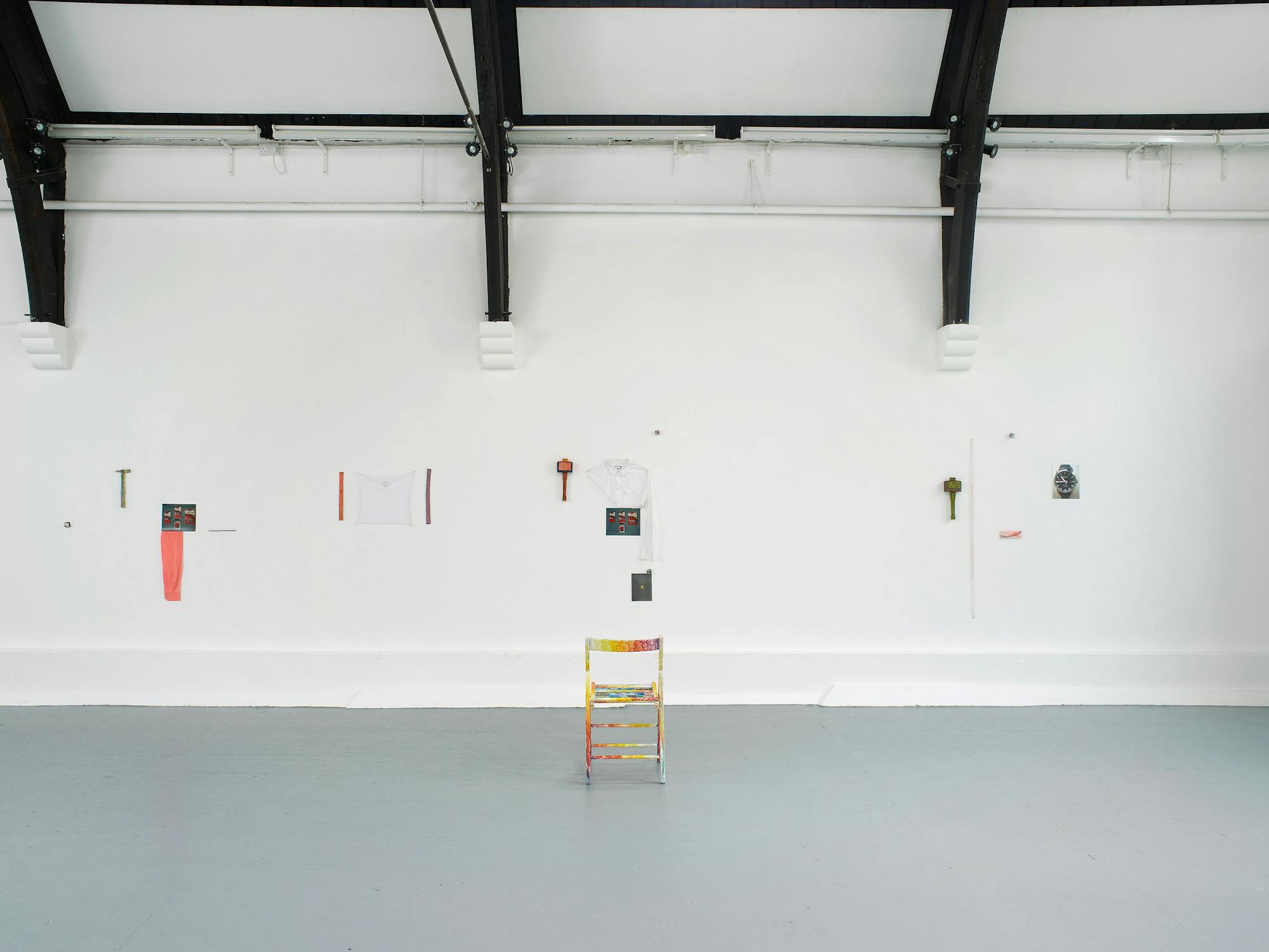 A minimalist art gallery with white walls displays small, spaced-out artworks and objects. At the centre, a colourful, painted chair faces the wall. Above and around it hang various items, including tools, papers, and small sculptural pieces, all evenly distributed.