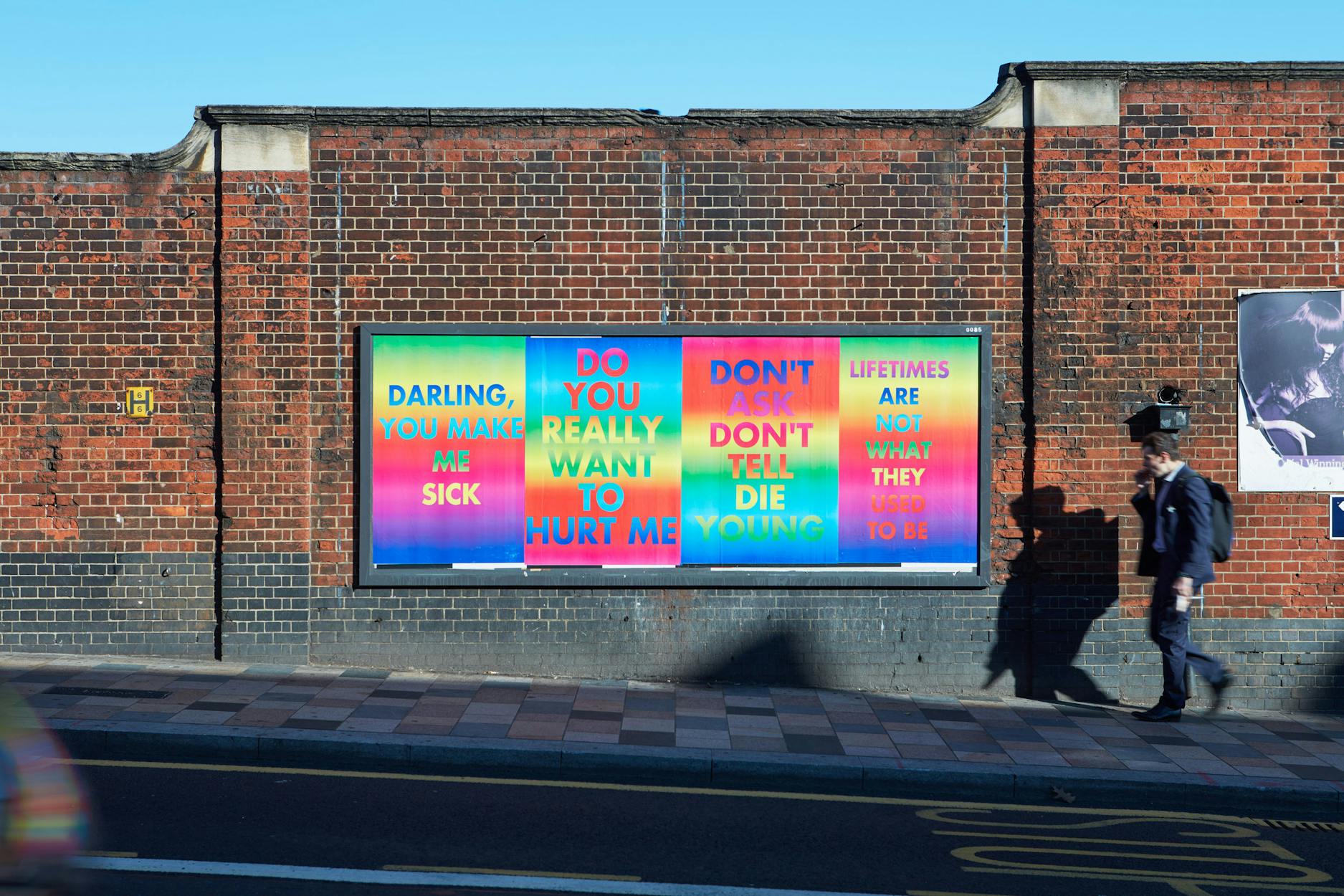 A colourful billboard campaign featuring four posters by David McDiarmid. Pasted on a brick wall. Bold text reads “Darling, you make me sick,” “Do you really want to hurt me,” “Don’t ask, don’t tell, die young,” and “Lifetimes are not what they used to be.”.