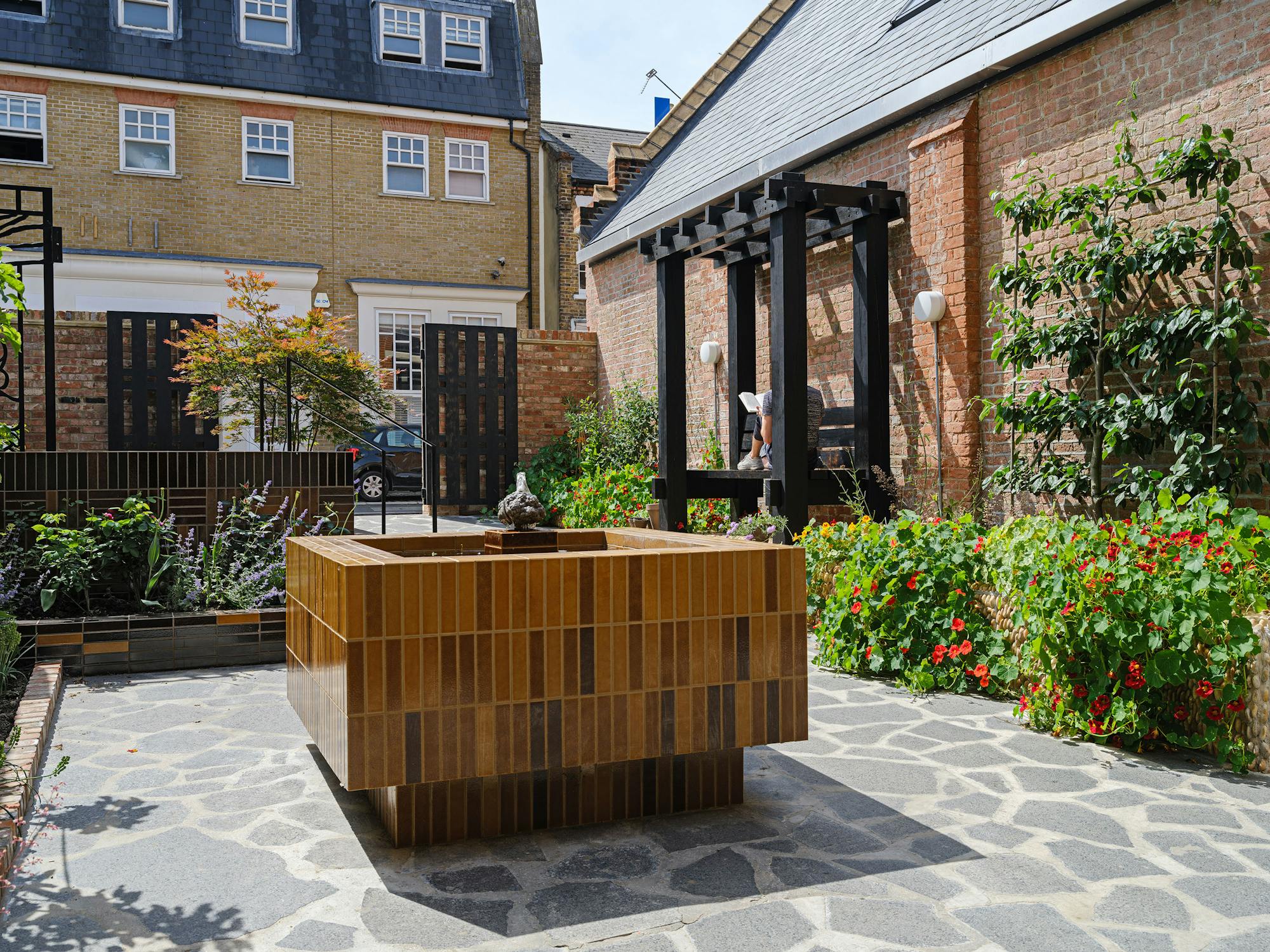 A courtyard garden features a rectangular water fountain covered in brown tiles at its centre. Surrounding the fountain are raised garden beds with various plants and flowers. In the background, there are brick buildings and a pergola with seating.