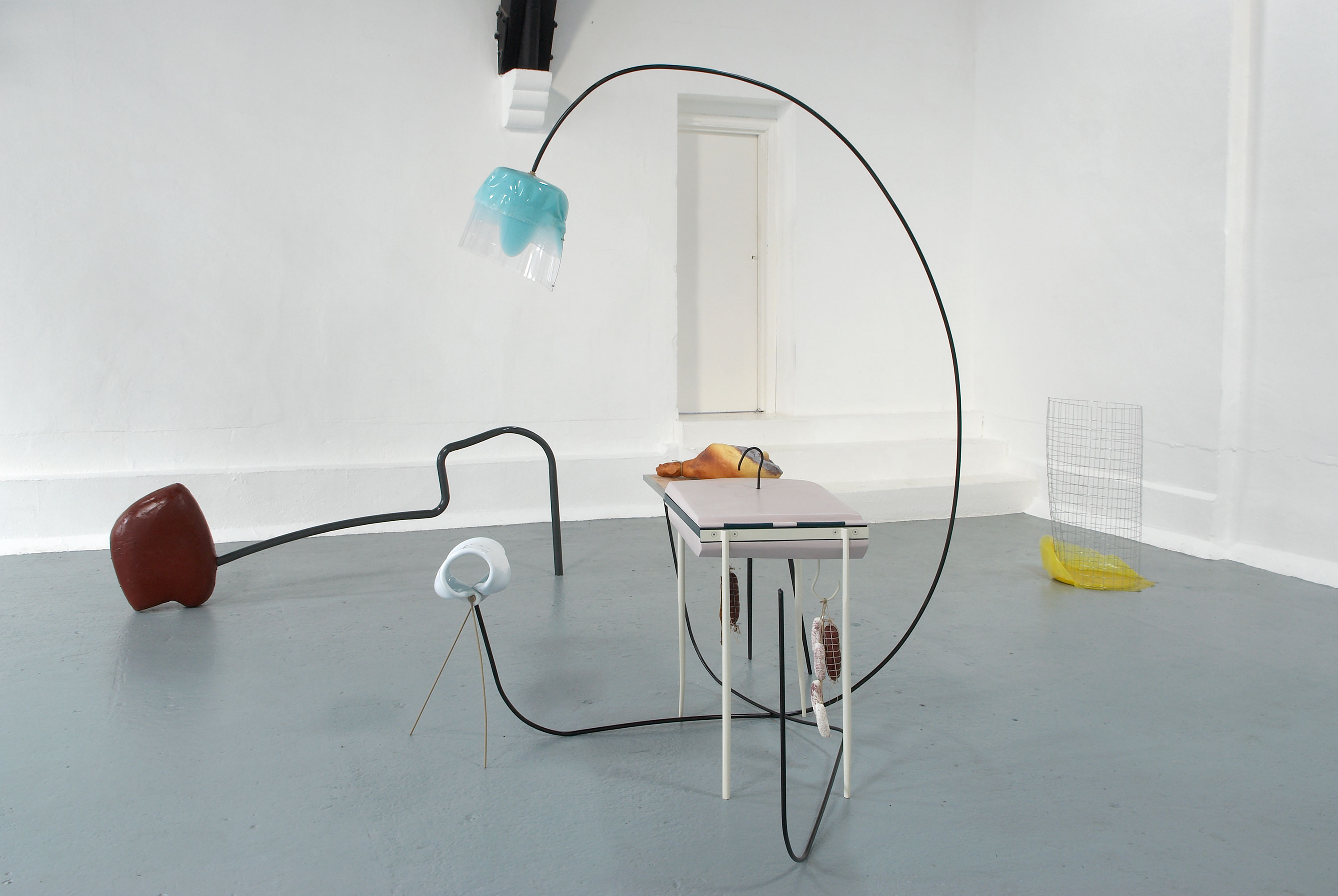 A minimalist art installation with two brown metal rods extending from the floor, each topped with smooth, abstract forms—one beige and one light grey. The setup stands against a white wall on a light grey floor.