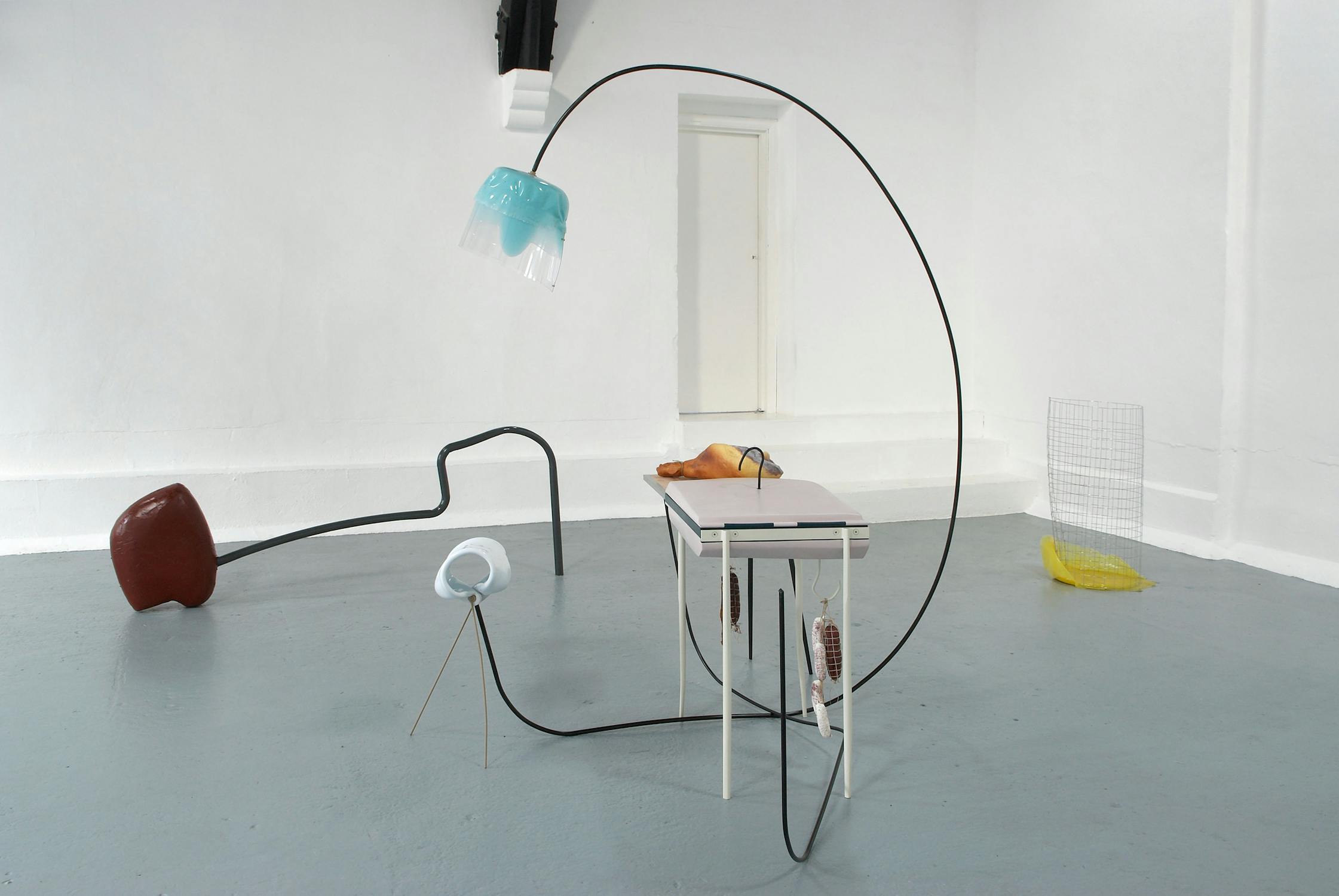 A minimalist art installation with two brown metal rods extending from the floor, each topped with smooth, abstract forms—one beige and one light grey. The setup stands against a white wall on a light grey floor.