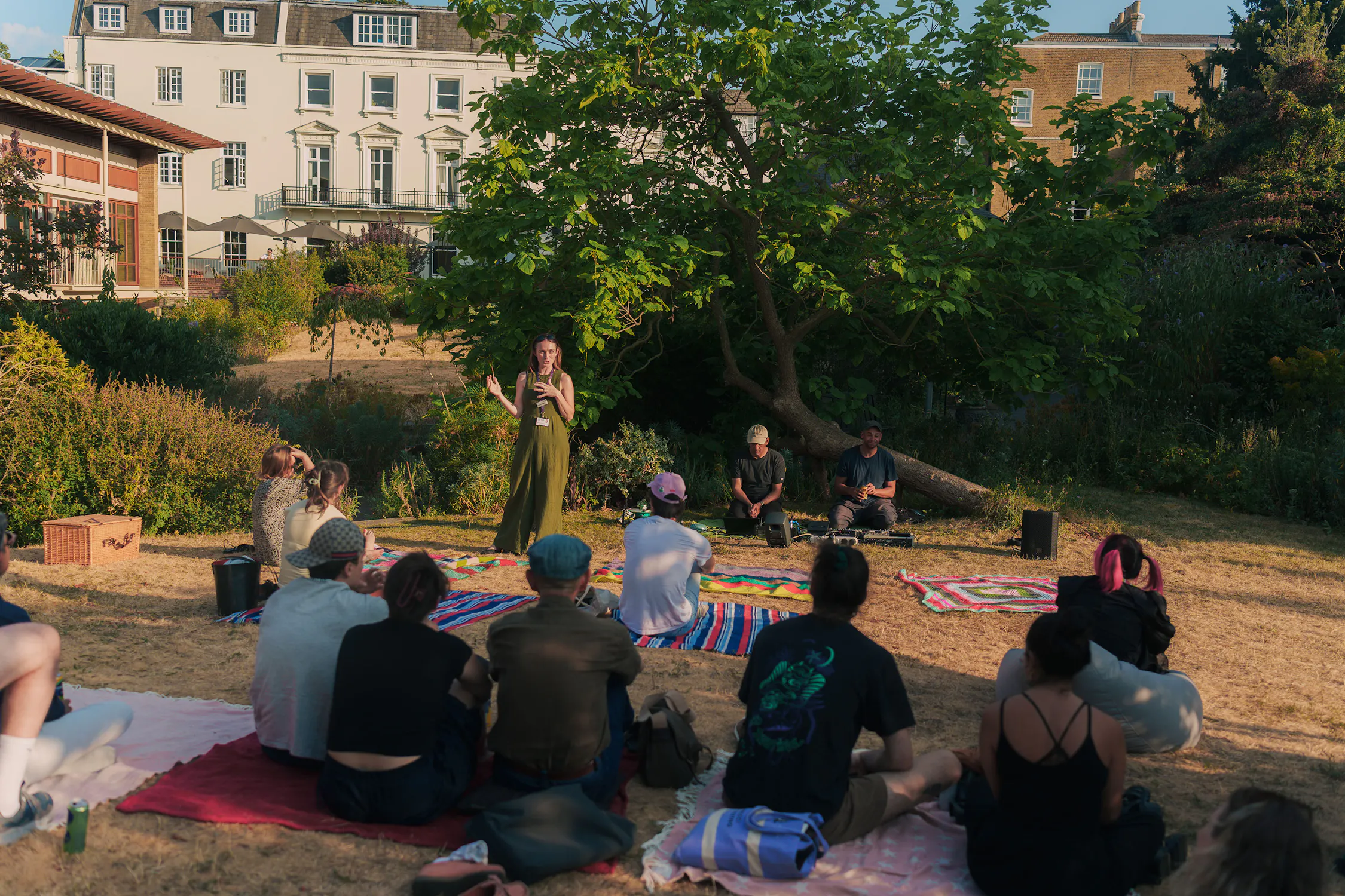 A person in a green boiler suit stands and speaks to a group of people sitting on blankets in a park. Behind them, a large tree provides shade, with white residential buildings visible in the background.