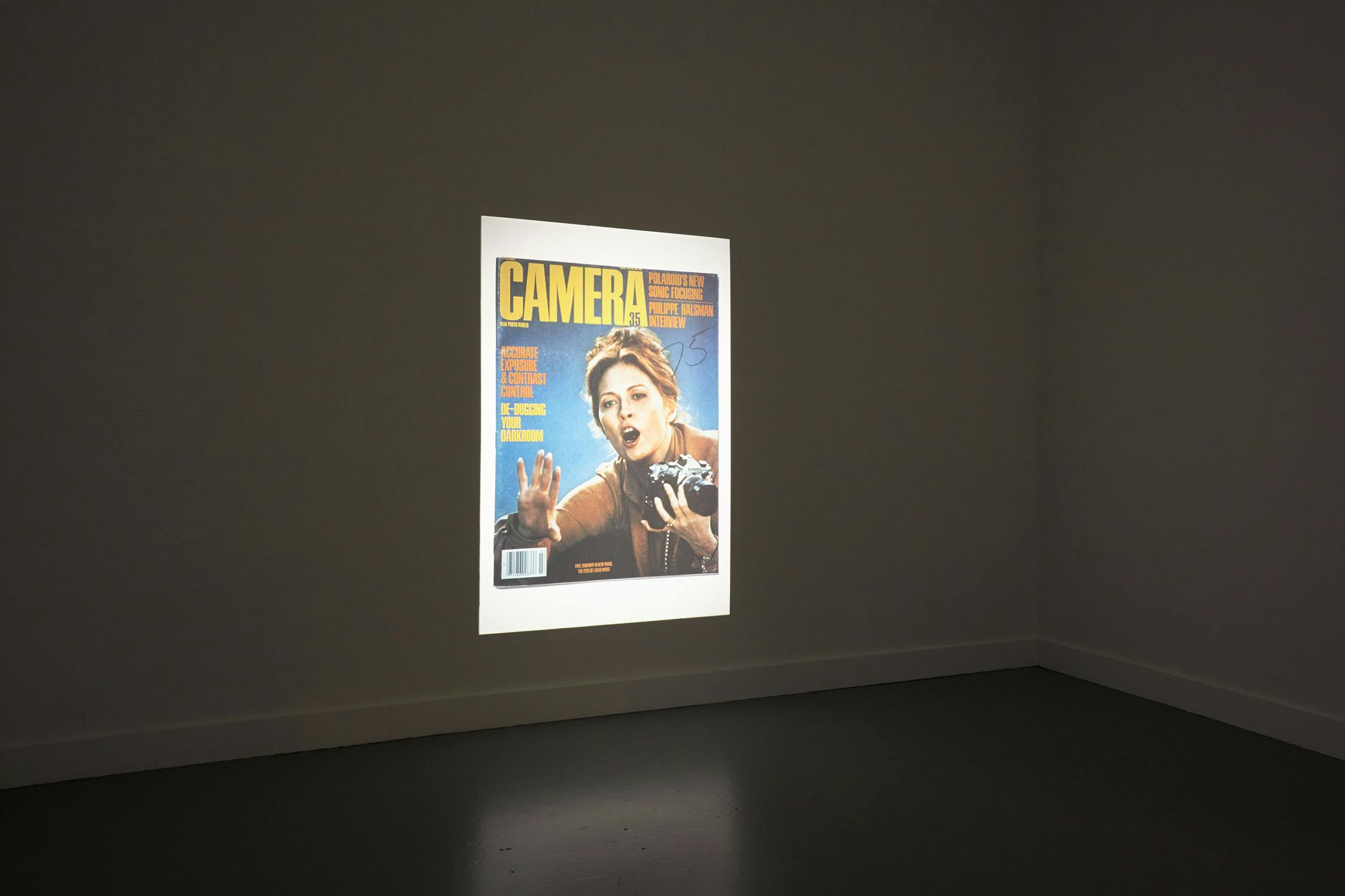 A photograph of a magazine cover titled "Camera" is projected onto a plain, grey wall. The cover features an expressive person holding a camera, with various headlines surrounding the image. The room is minimalistic, with no additional objects or decorations.