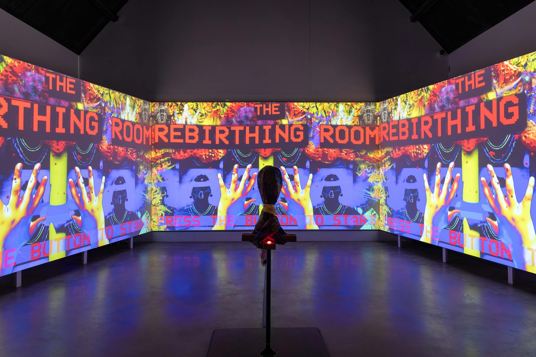 a photograph of three screens that are projected with bright and graphic images. There is text that reads 'THE REBIRTHING ROOM'. In the foreground there is a hooded object on a stand. 