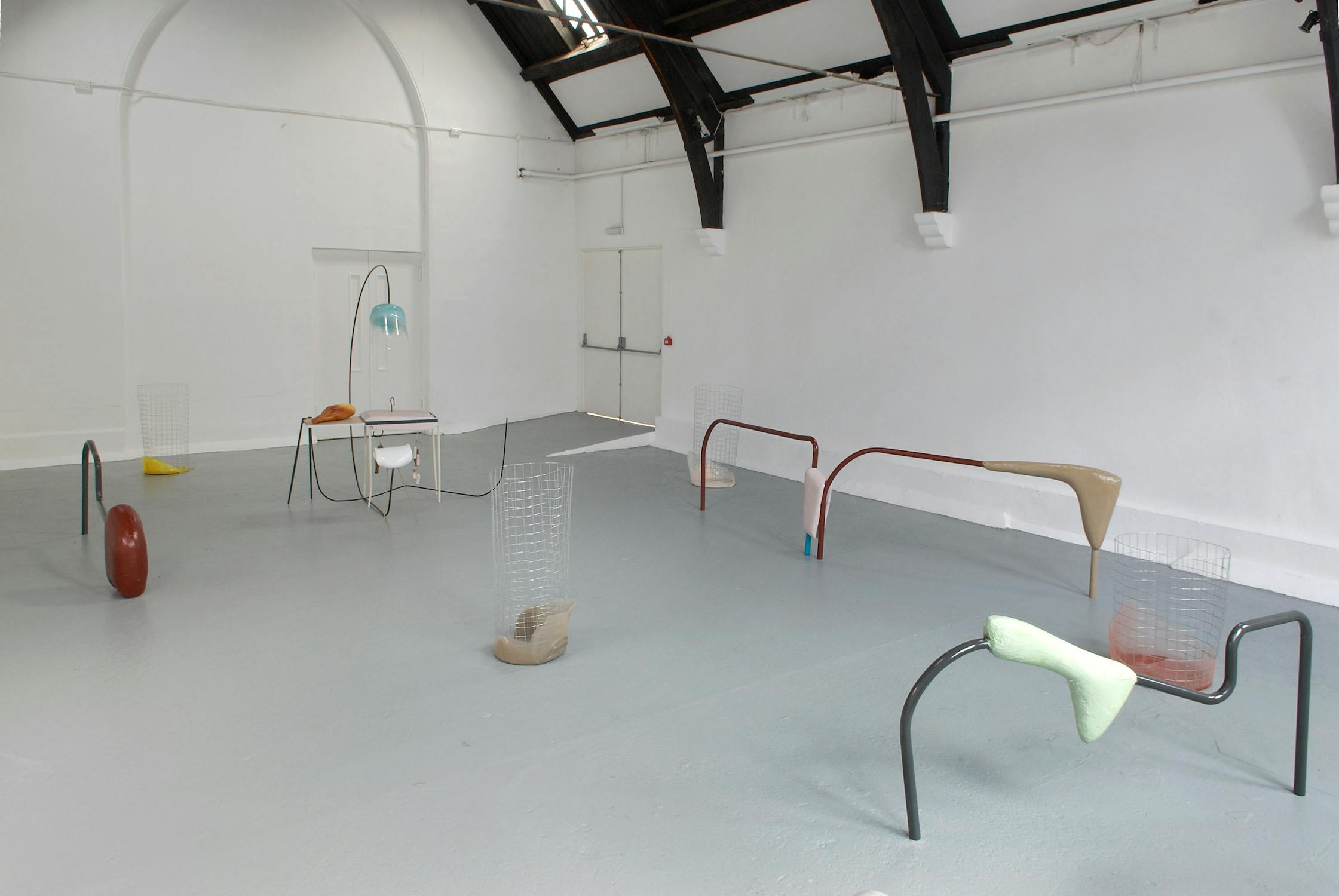 A white walled room with a grey floor features an art installation comprised of various abstract sculptures. The pieces include metal rods, mesh, and colorful shapes, arranged in a spacious setting with natural lighting from above.