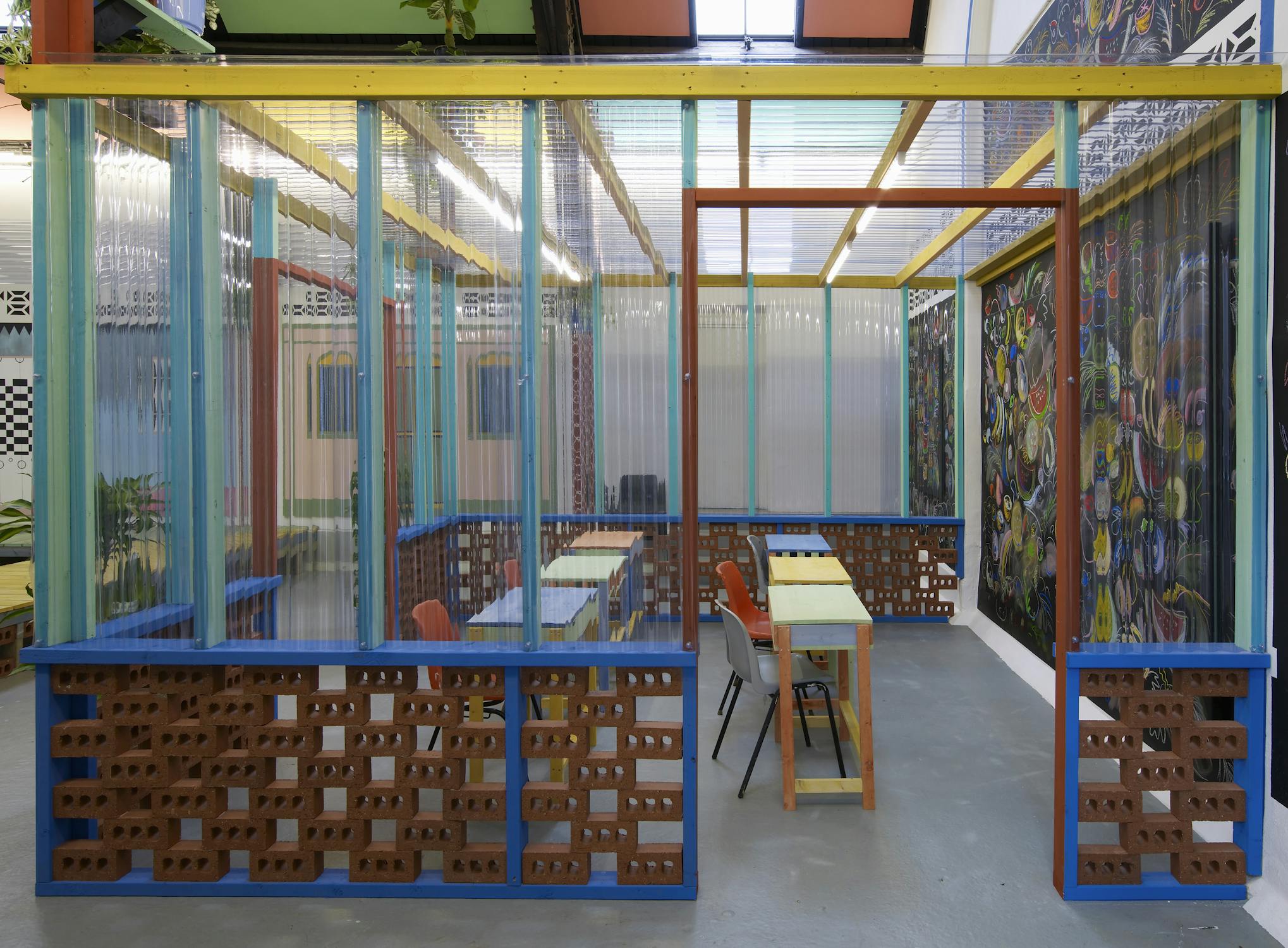 Part of Sol Calero’s exhibition at Studio Voltaire, this enclosed area includes orange chairs and tables set within a semi-enclosed structure made of clear corrugated plastic, blue framing, and decorative brickwork. Surrounding walls are painted with vivid murals that evoke tropical and domestic themes.