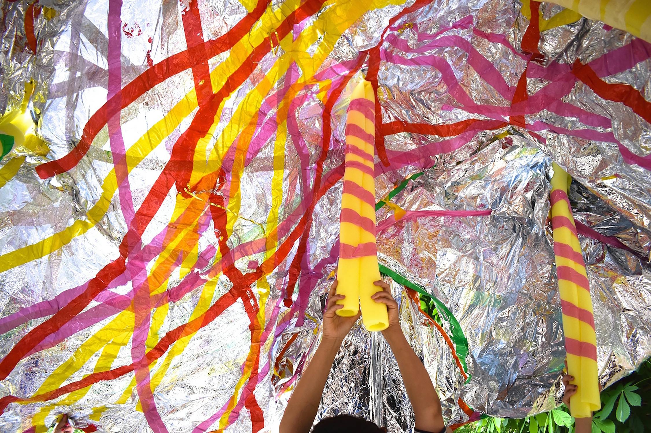 A child reaches up towards two large, colorful candles hanging under a vibrant, shiny canopy decorated with pink, red, and yellow streamers. The scene is festive and bright, with the sunlight reflecting off the metallic surfaces.