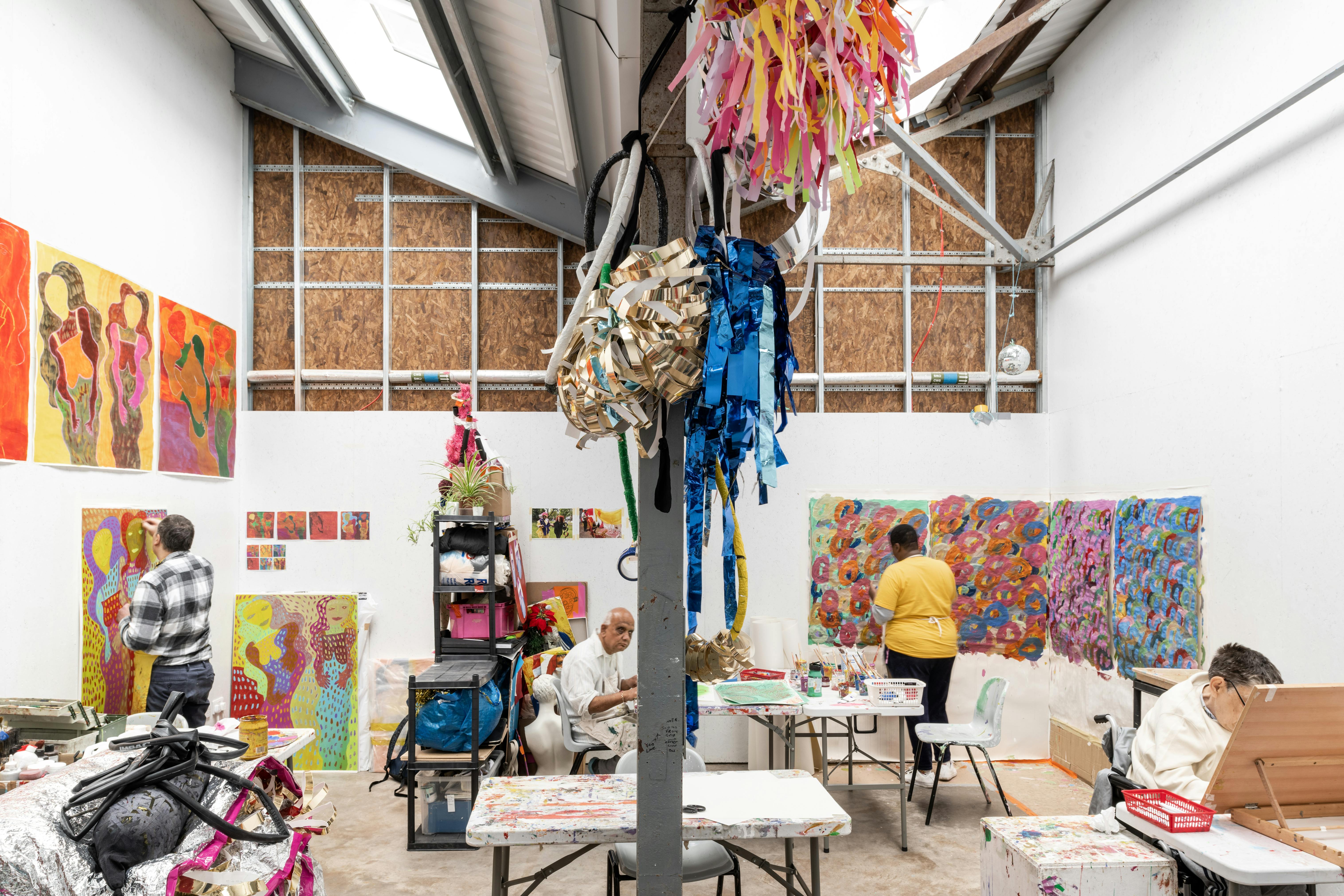 A bright art studio with high ceilings, tables, and art supplies. Several people are painting or drawing. Colorful artworks and murals cover the walls. Hanging decorations and ribbons add to the lively, creative atmosphere. Natural light streams in from above.