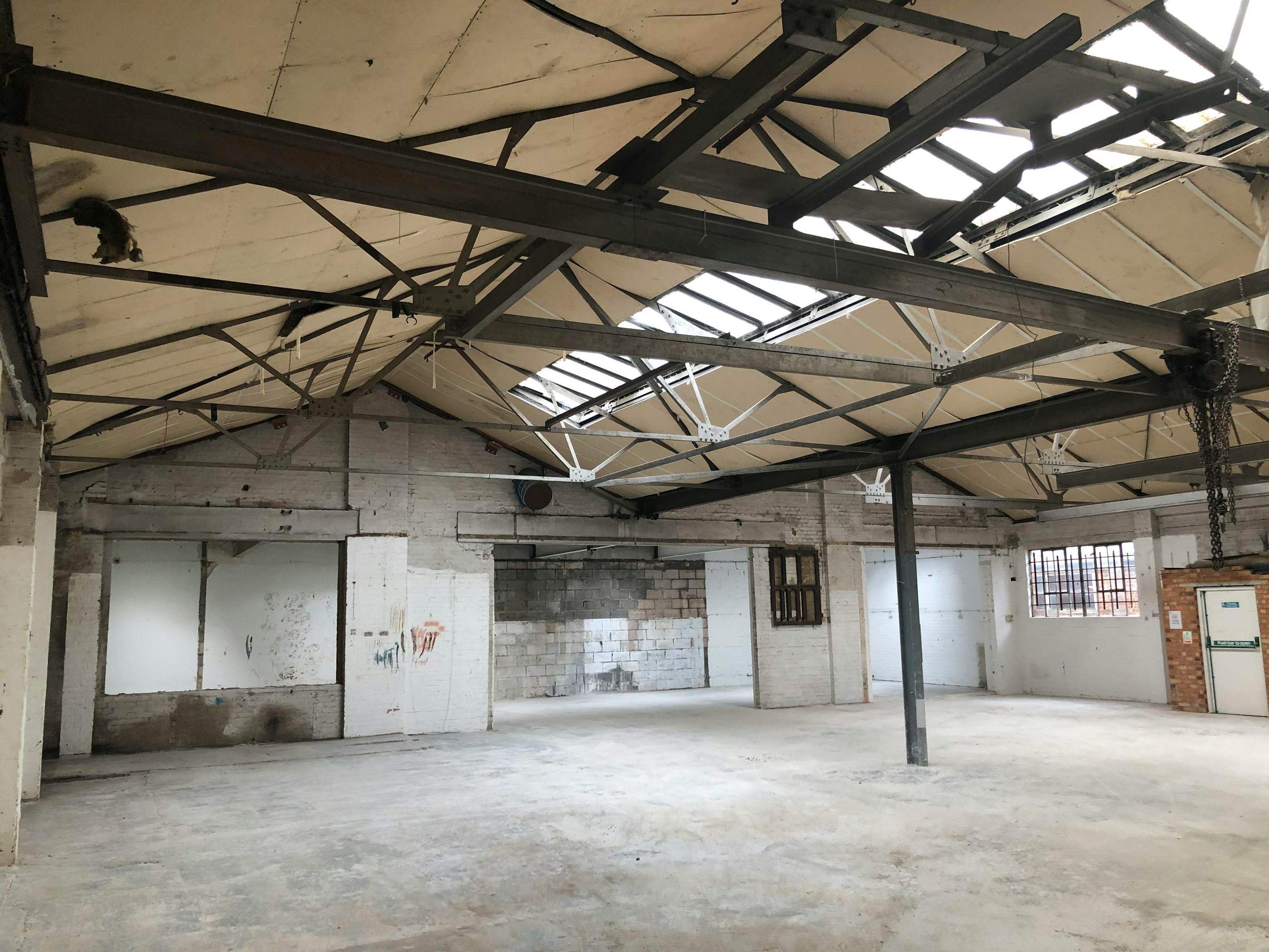 Large, empty industrial warehouse with exposed metal beams, skylights and peeling white walls. The floor is bare concrete, and there are windows and large doors on the far wall. Natural light fills the space.