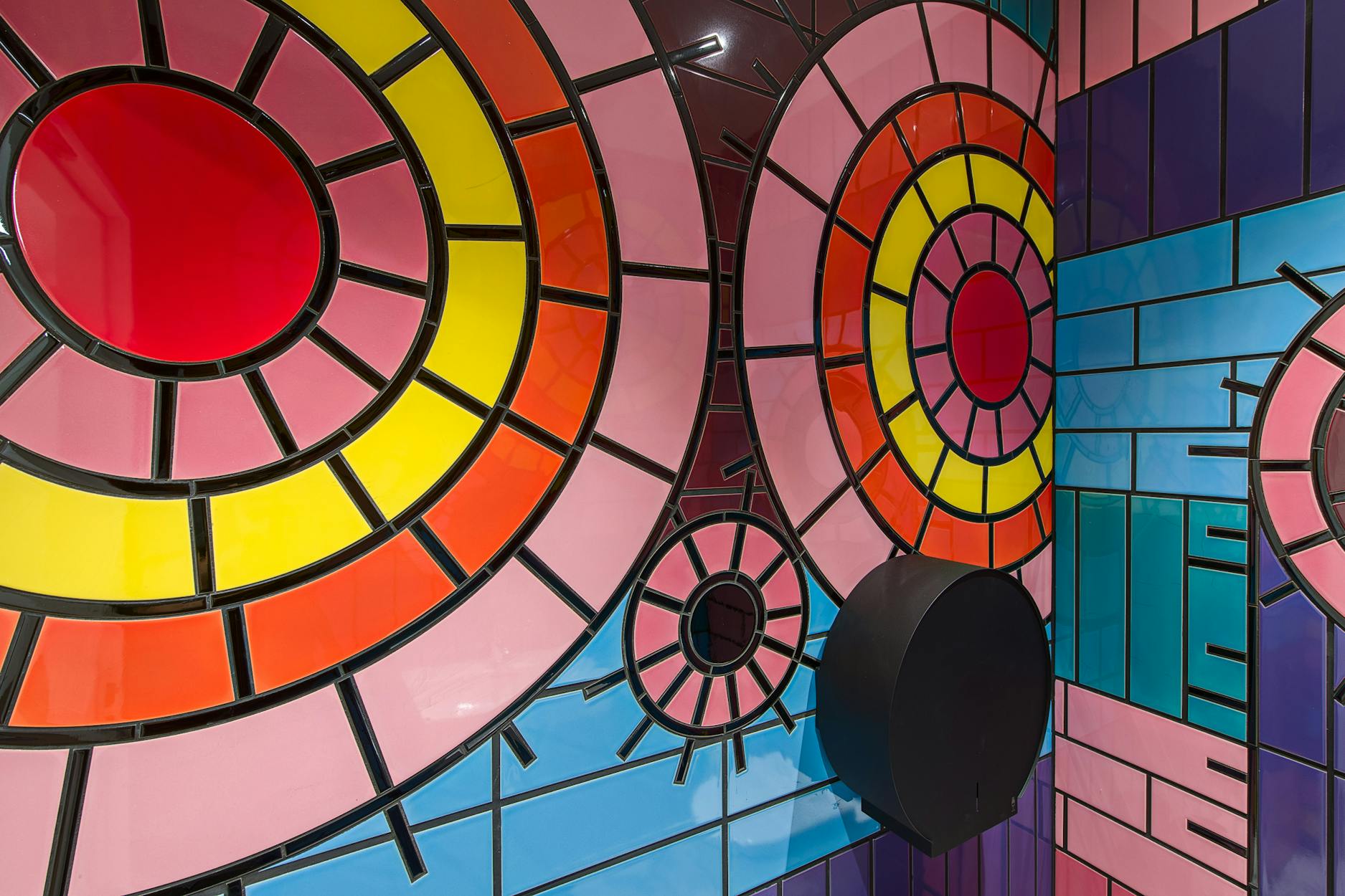 A vibrant tiled bathroom wall featuring abstract geometric designs. Large circular patterns in bold colours such as red, yellow, pink, and orange dominate the artwork. Black lines separate the colourful sections, creating a stained glass effect.
