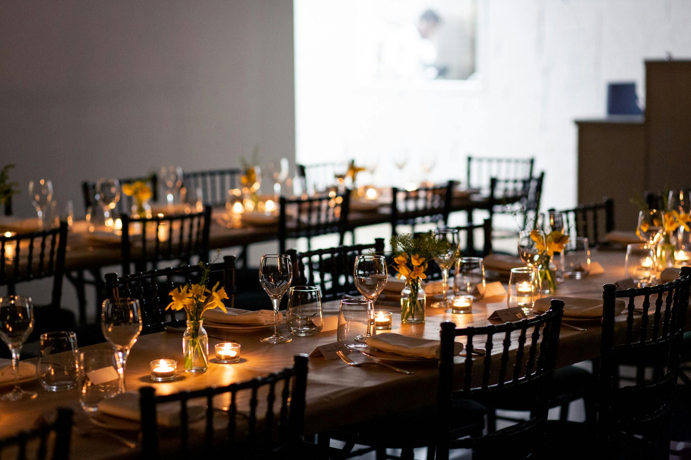 A soft focus photograph of two long tables set for dinner. The room is dark and the tables are light by small candles.