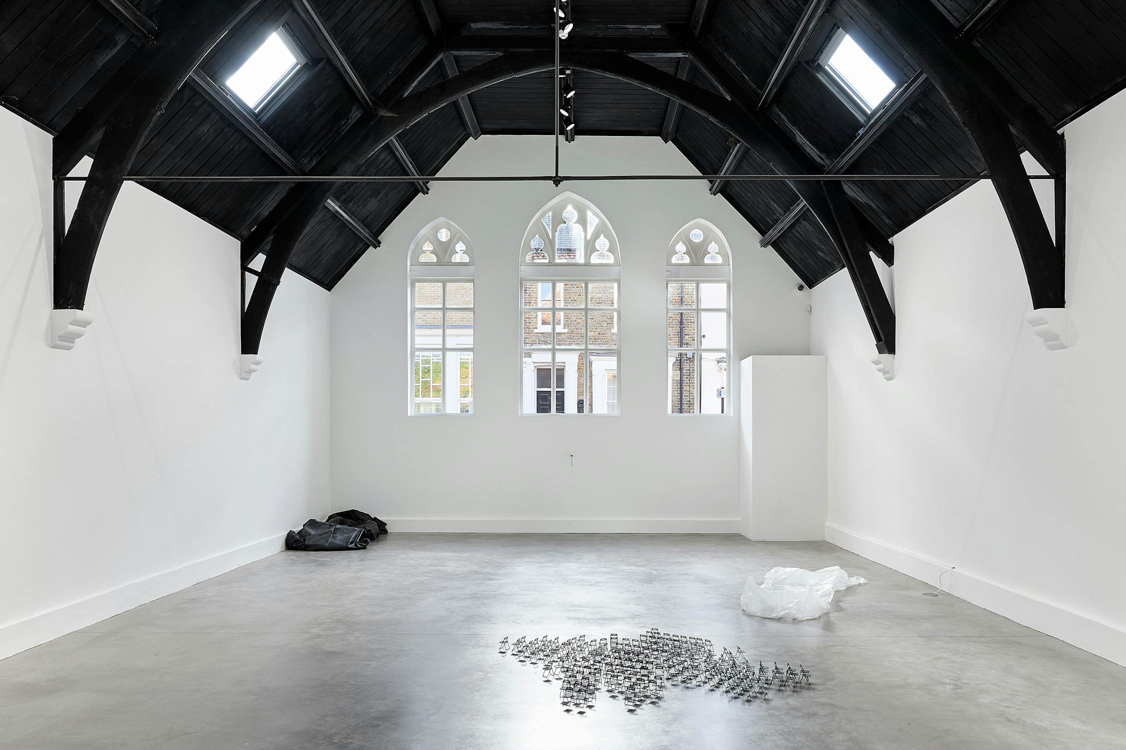 An art gallery with white walls, a high arched black ceiling, large gothic-style windows, and a polished concrete floor. Minimalist sculptures by Solomon Garçon and materials are arranged on the floor near the windows.