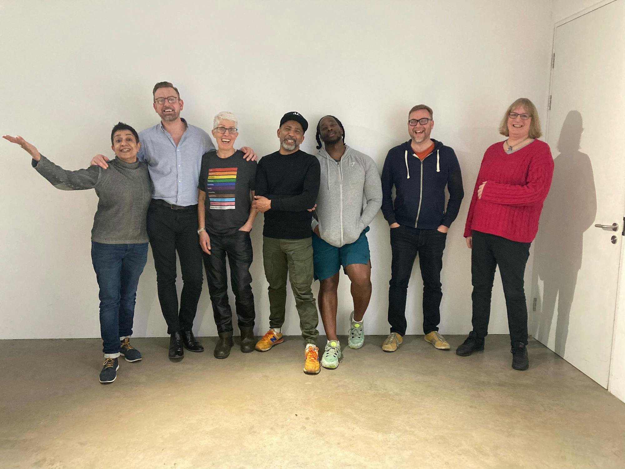 Seven people stand side by side in a well-lit room against a plain wall, smiling and posing casually together. They appear relaxed and happy, dressed in casual clothing.