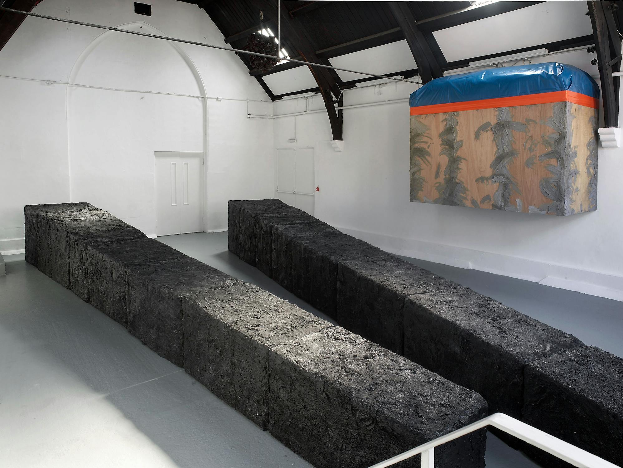 A white gallery with tall ceilings features two long, rectangular, black textured blocks on the floor. On the wall, a large box wrapped in plastic with painted patterns and a blue tarpaulin on top is suspended above the ground.
