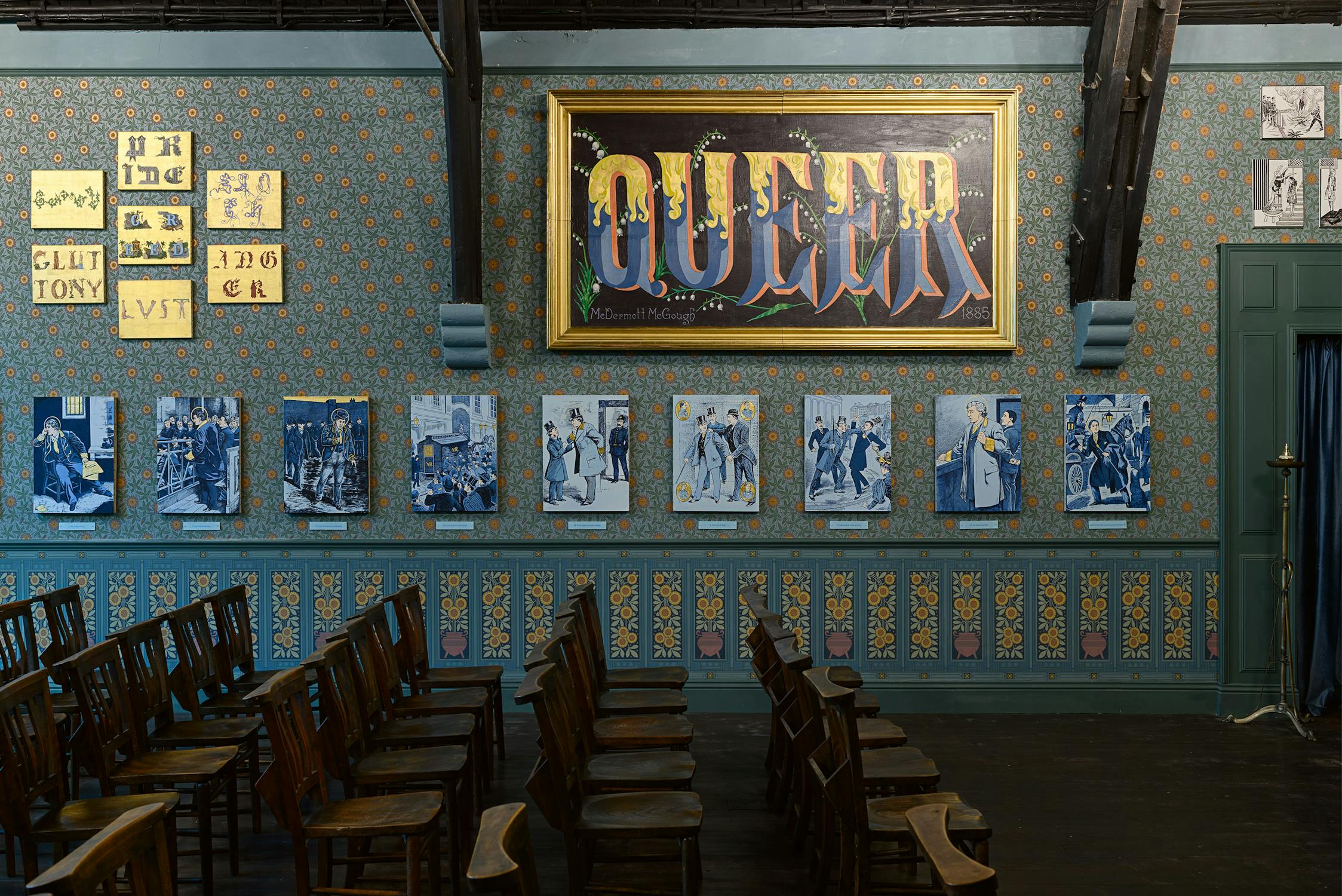A room with ornate floral wallpaper, wooden chairs facing forwards, and framed blue-toned portraits on the wall. Above them hangs a large gold-framed artwork with the word “QUEER” in bold letters. Smaller yellow and black artworks are to the left.