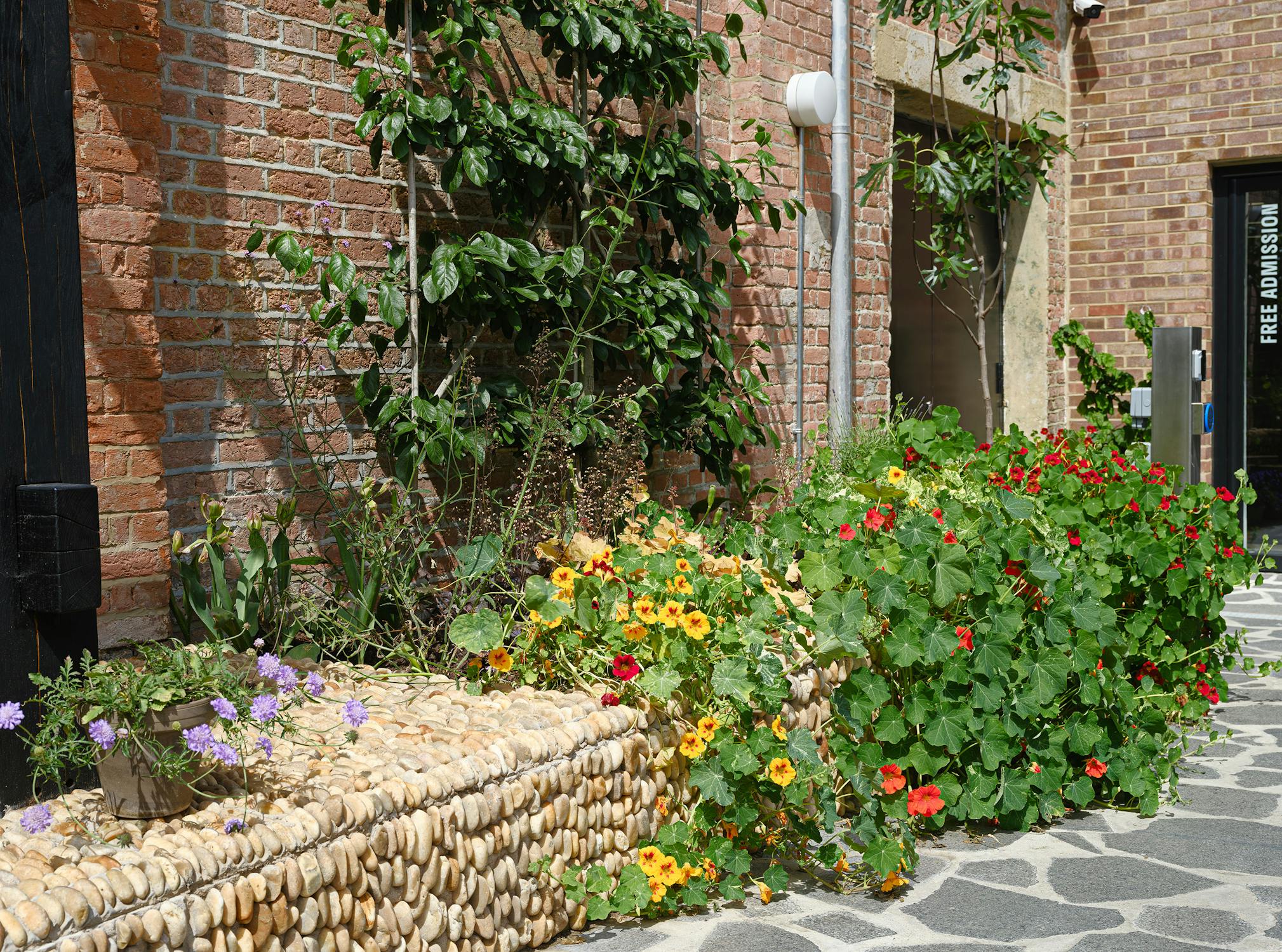 A brick-walled building entrance surrounded by vibrant flower beds and greenery. The door has signs reading "ART EVENTS CAFÉ SHOP" and "FREE ADMISSION ALL WELCOME." The path leading to the entrance is paved with irregular stone slabs.