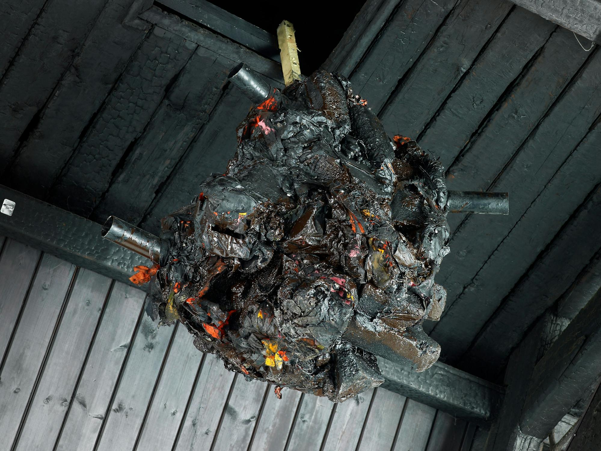 A large, dark, irregularly shaped sculpture hangs from a rope beneath a wooden ceiling. The sculpture has rough, jagged textures, metallic pipes protruding from it, and glossy black with hints of red, orange and silver that suggest molten or charred materials.
