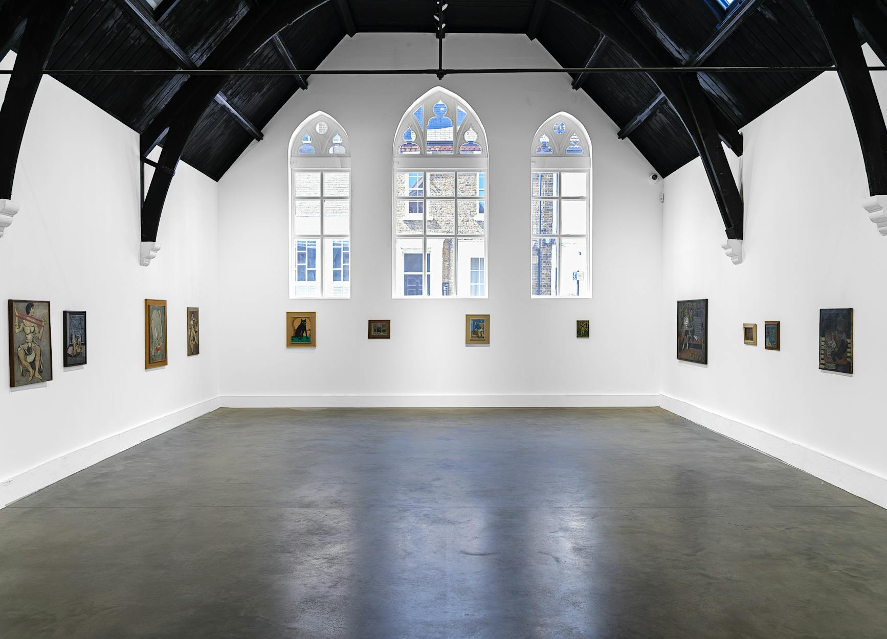 A bright, spacious art gallery with high, dark vaulted ceilings and tall arched windows. Various framed paintings are displayed on the white walls around the room. The gallery floor is bare and grey.