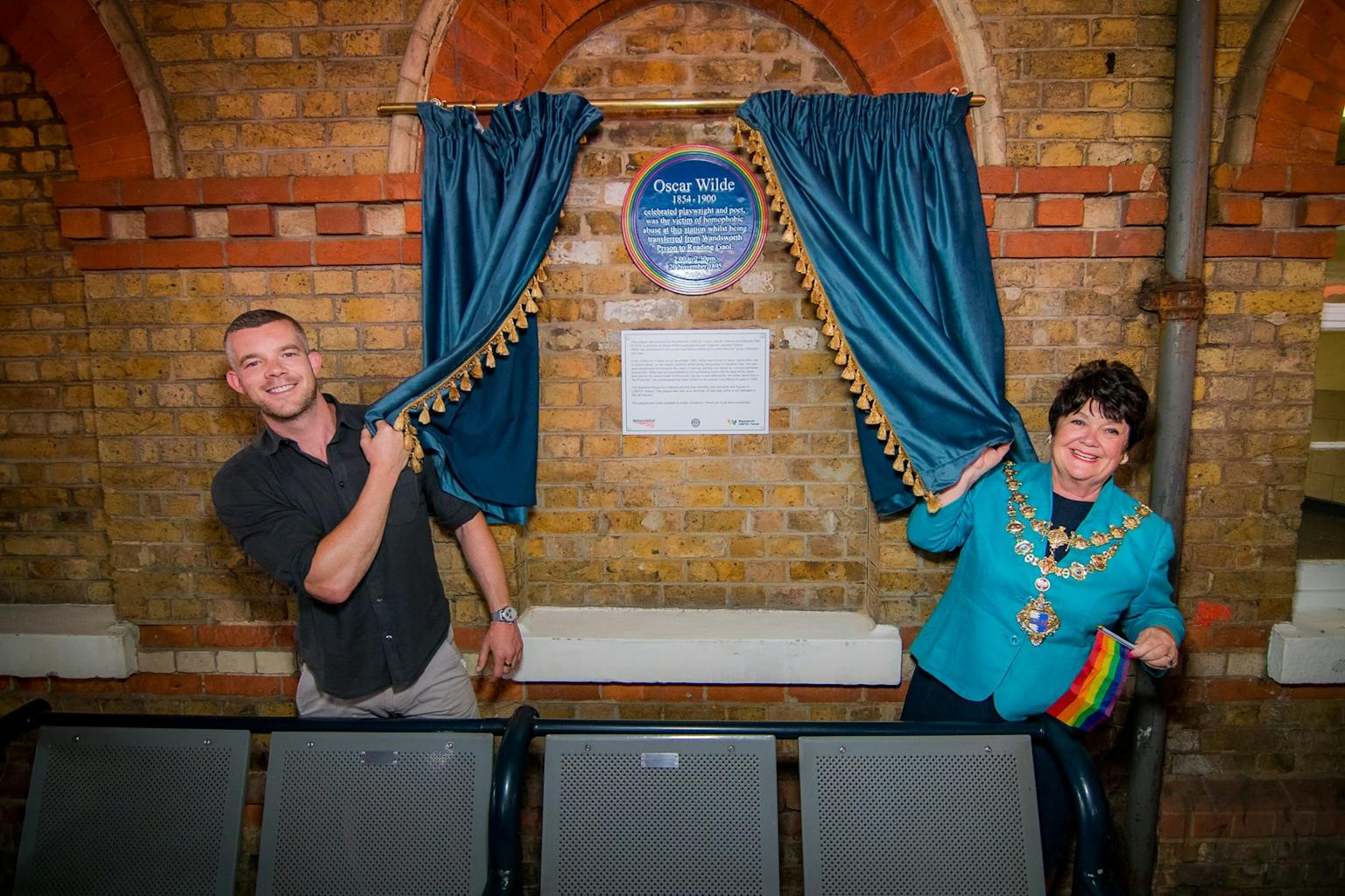A man and a woman unveil a blue plaque dedicated to Oscar Wilde on a brick wall. The woman wears a ceremonial chain and holds a rainbow flag, while both smile toward the camera. Blue curtains frame the plaque.