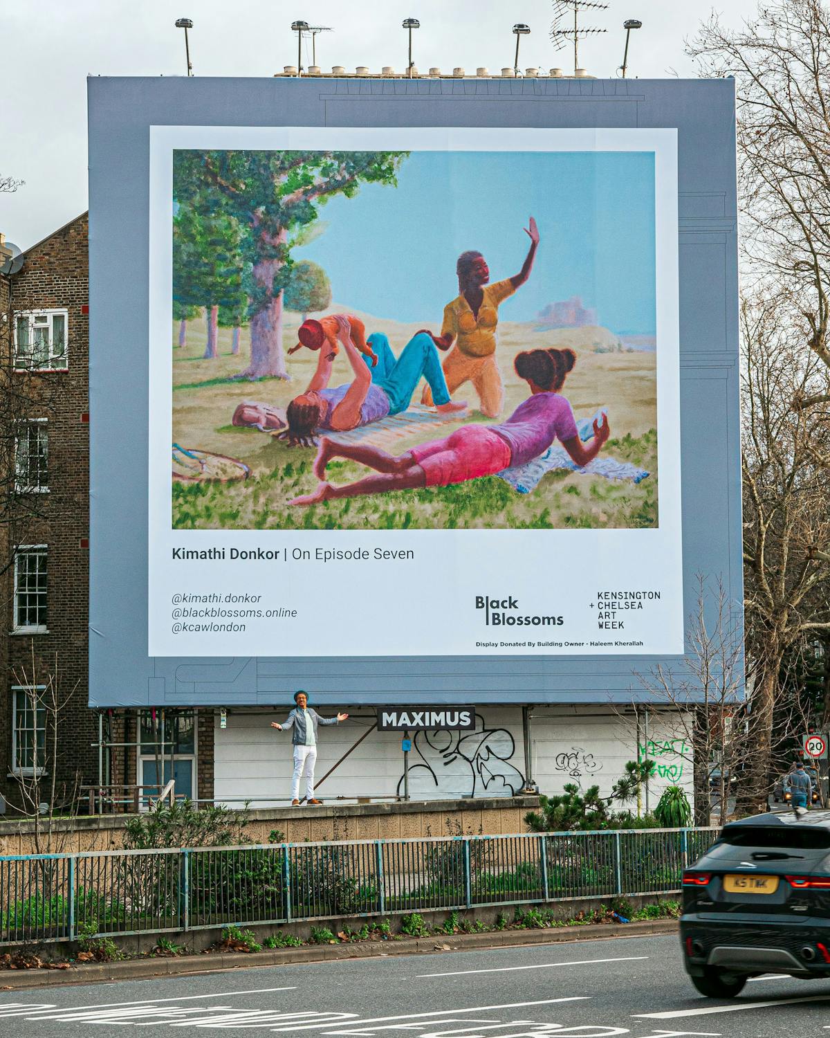 A large billboard displays a colourful painting of four Black women relaxing on grass with a coastal background. Below the art, text credits Kimathi Donkor and Black Blossoms. A person stands below; nearby, cars pass on a tree-lined street.