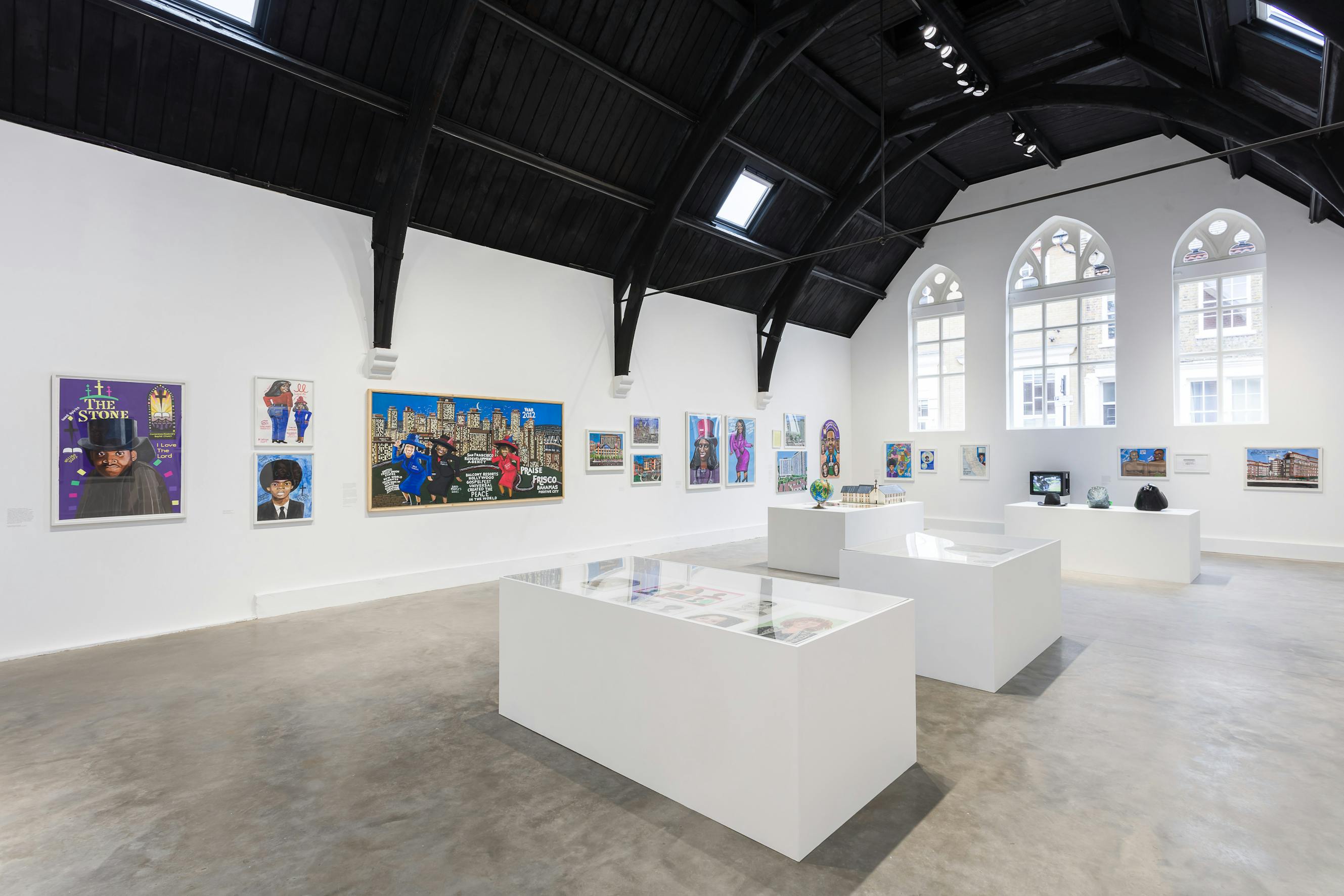 a gallery with white walls and a black vaulted roof. In the middle of the room there are square vitrines with glass tops. Artworks of various painted public figures are mounted on the walls in the background.