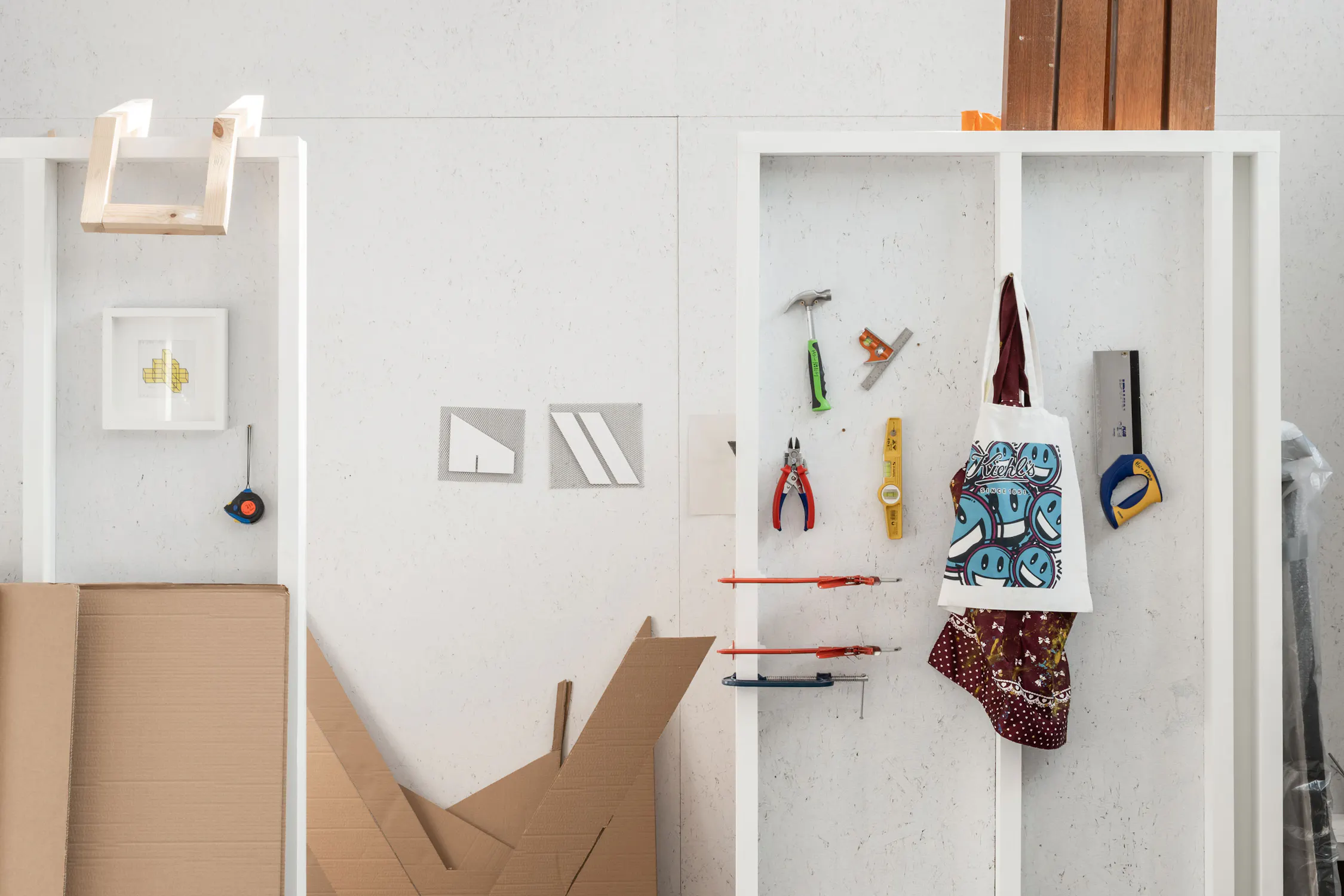 A white shelving unit displays hanging tools, a printed tote bag, and carpentry items. Cardboard pieces and stacked wooden planks lean against the wall. Framed art and geometric paper are also visible, creating a tidy, organised workspace.