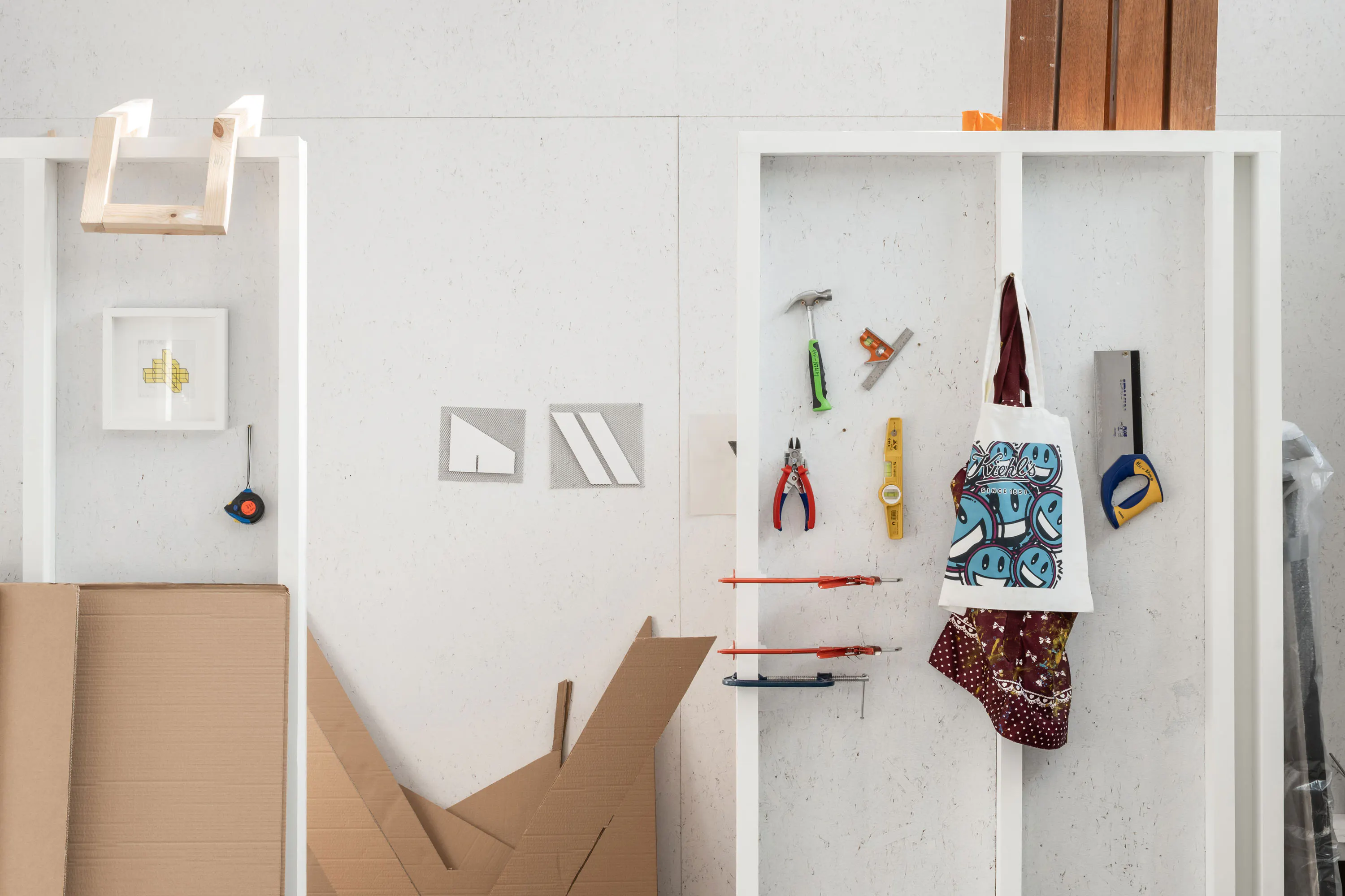 A white shelving unit displays hanging tools, a printed tote bag, and carpentry items. Cardboard pieces and stacked wooden planks lean against the wall. Framed art and geometric paper are also visible, creating a tidy, organised workspace.