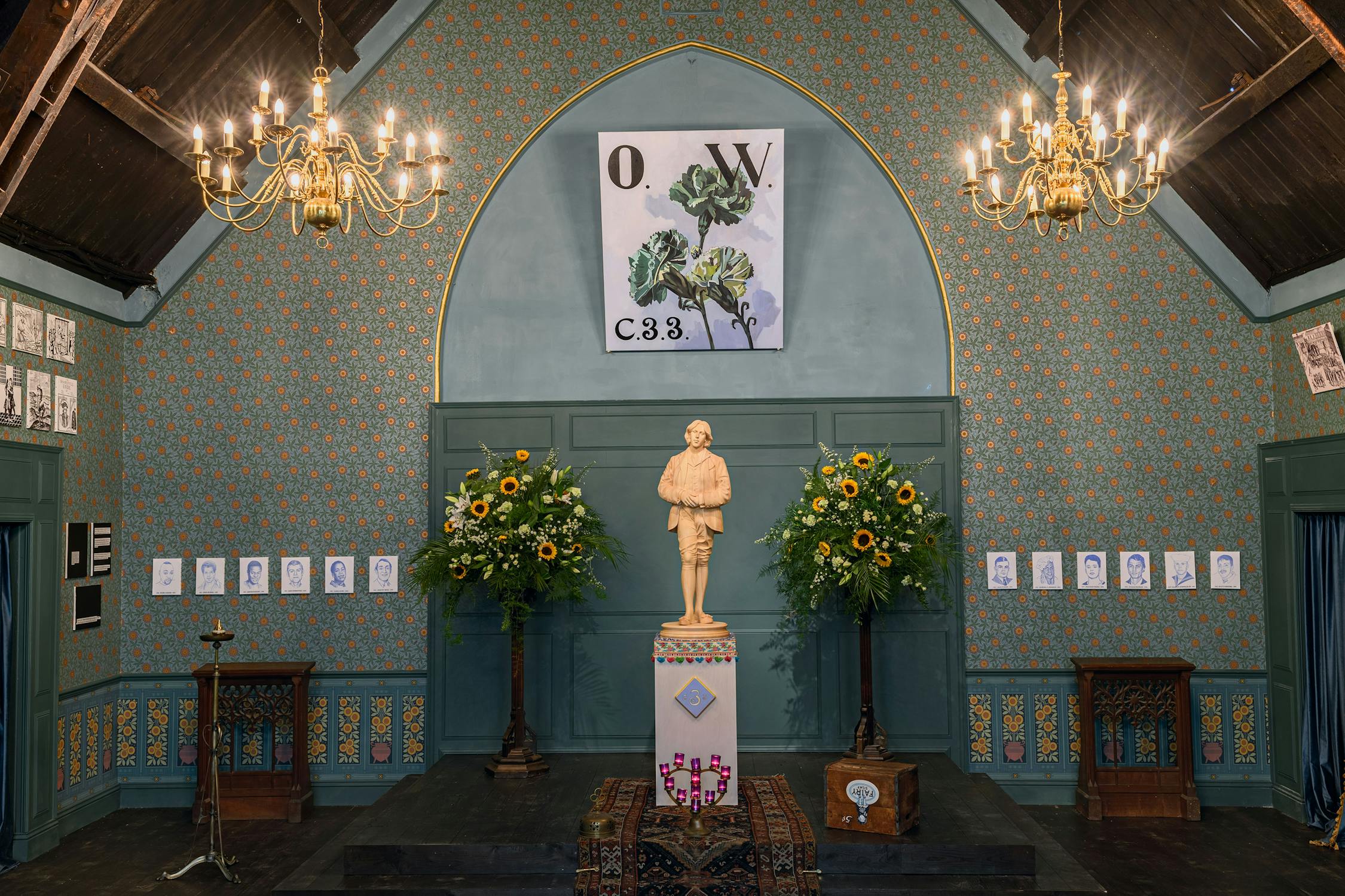 An ornate room with dark wood ceiling, patterned wallpaper, and chandeliers. A statue of a person stands on a decorated plinth between two large floral arrangements. Above is a painting with "O.W. C3.3" and green leaves. Portrait drawings line the walls.
