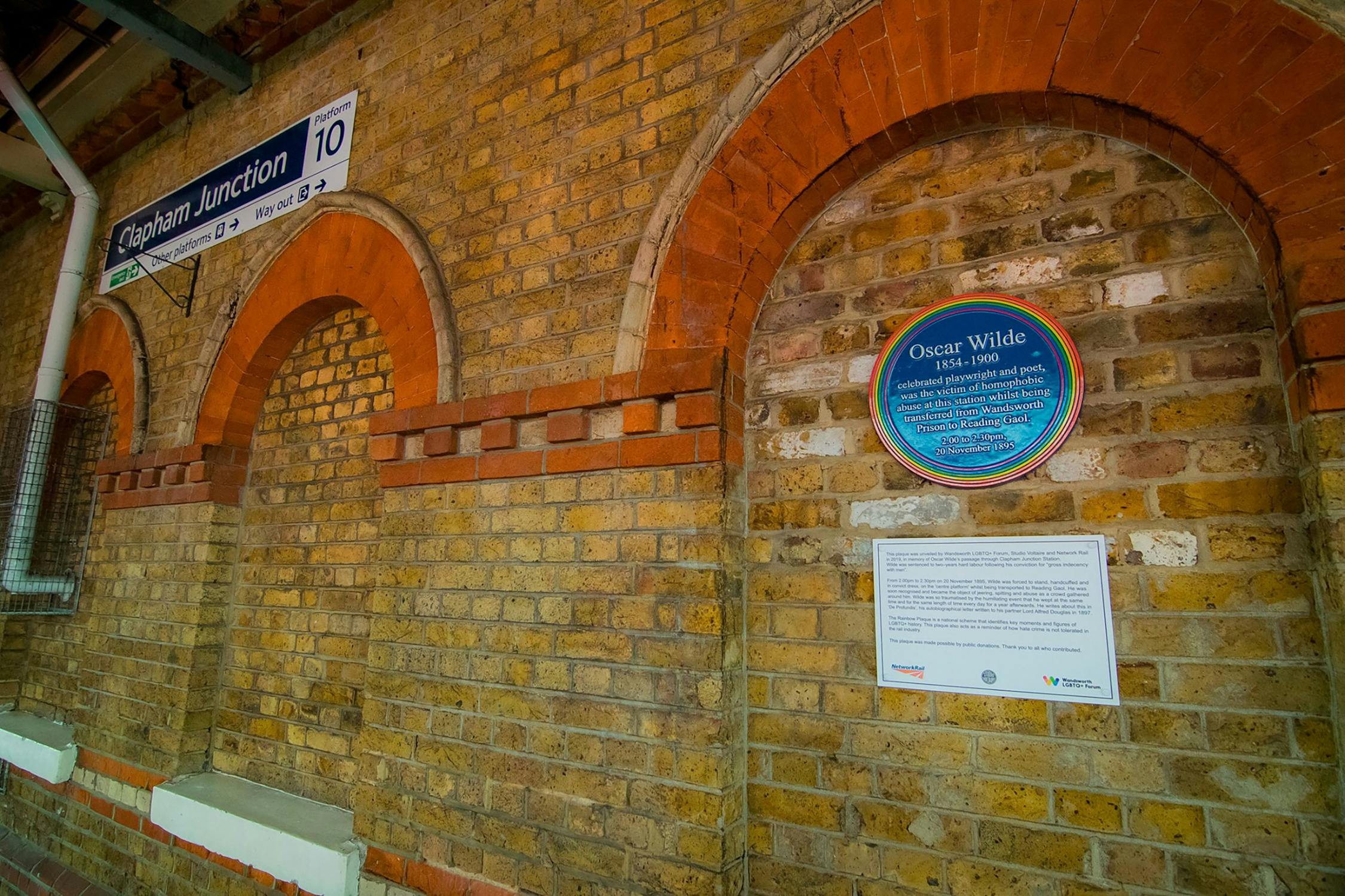 A brick wall at Clapham Junction station with a blue plaque honouring Oscar Wilde, and an accompanying information sign. The station sign and arched brickwork are clearly visible.