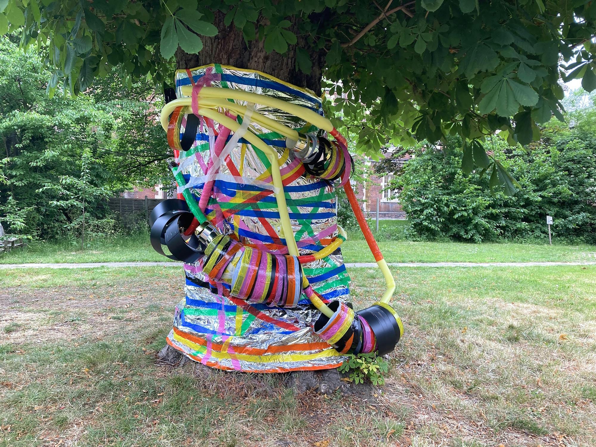 A tree trunk wrapped in colorful tapes and decorated with hula hoops and foam tubes. The vibrant installation contrasts with the green park setting in the background.
