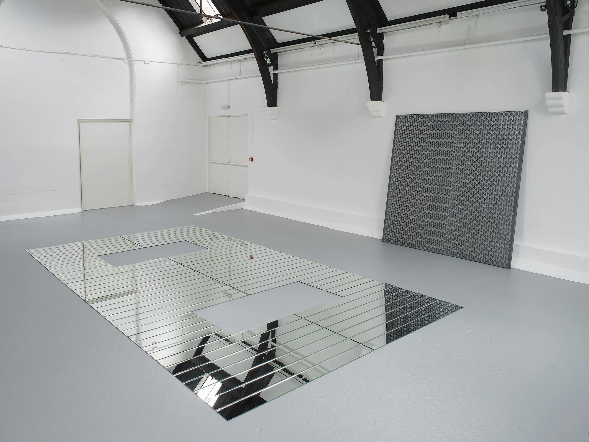 A minimalist white gallery room features a large, rectangular mirrored floor piece with geometric cut-outs. Against the back wall leans a square panel covered in a grid of dark circular shapes. Exposed black beams arch along the ceiling.